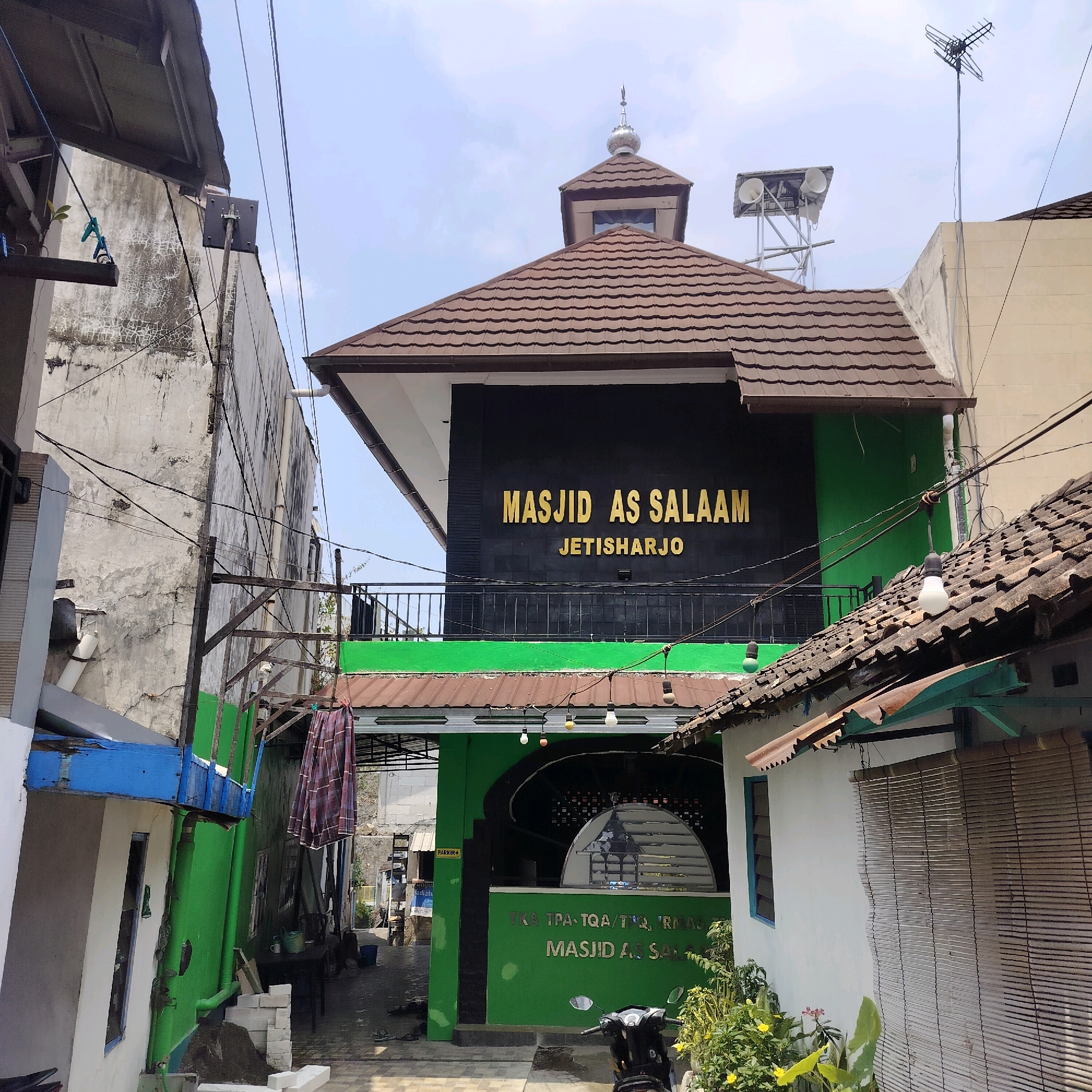 The image shows the Masjid As Salaam in Jetisharjo, Yogyakarta, Indonesia. The mosque is a two-story building with a green facade on the lower level and a black facade on the upper level featuring the mosque's name in gold lettering. The mosque has a brown tiled roof and a small dome topped with a crescent moon symbol. In front of the mosque, there is a street with a parked motorcycle. Several power lines crisscross the scene, and there are buildings on either side of the mosque. The sky is partly cloudy. The area appears to be a mix of residential and commercial structures, typical of a bustling urban environment in Yogyakarta.