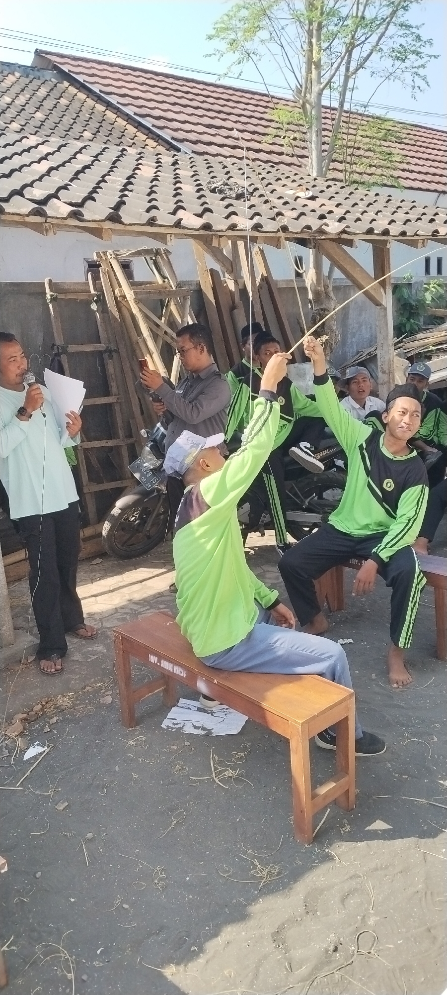 The image depicts an outdoor gathering in Jombang, Indonesia. In the foreground, two young men in bright green and black long-sleeved shirts are seated on wooden benches. One man on the right is holding a long, thin object, possibly a kite string or fishing rod, with his right arm extended upwards. The other man on the left is looking towards the right and has his left arm extended upwards. Behind them, several other young men are gathered, some wearing similar green and black attire, and one appears to be holding a mobile phone.
To the left, a man wearing a light blue long-sleeved shirt and black pants is speaking into a microphone and holding some papers. He is standing and facing towards the gathered individuals. To his right, a man in a grey long-sleeved shirt is looking down at his phone. A motorcycle is parked behind the group.
The background reveals a building with a tiled roof and wooden support structures. The ground appears to be dirt or sand, scattered with dry grass and debris. The sky is clear and blue, suggesting a sunny day. The overall impression is a casual outdoor event or activity.