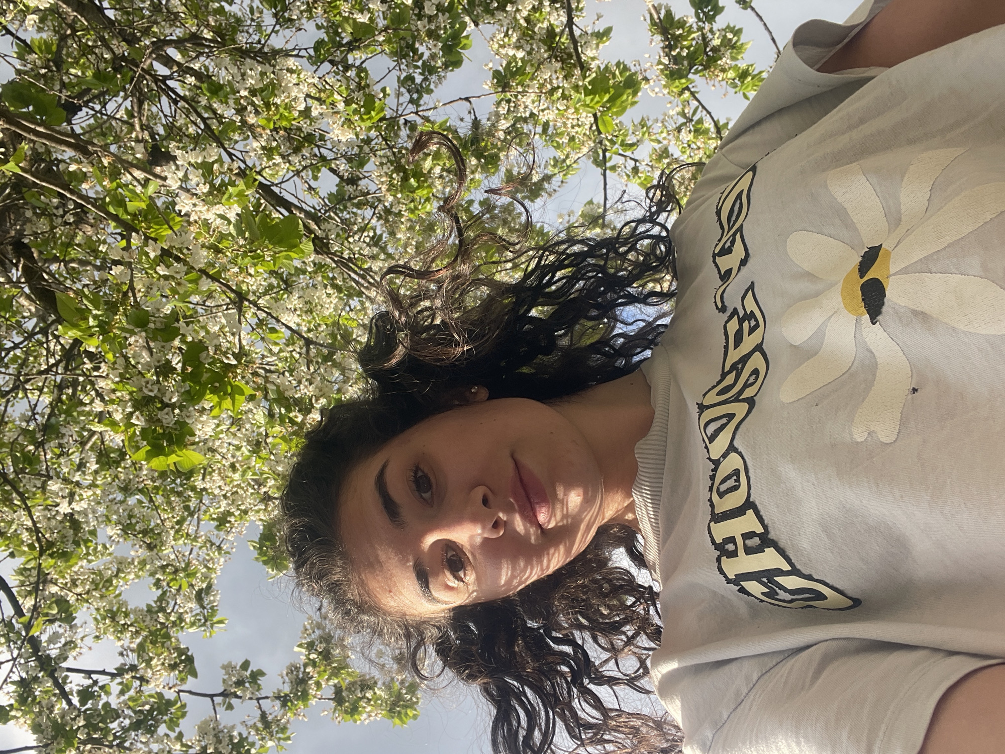 A low-angle portrait captures a young woman looking directly into the camera, which is positioned below her, creating a worm's-eye perspective. She has dark, curly hair and her face is partially shaded by dappled sunlight, creating patterns of light and shadow. Her expression is relaxed with a subtle, gentle smile.
She is wearing a light-colored t-shirt adorned with a large, stylized white daisy design with a yellow center. Below the daisy, or integrated into the design, the word "HOOD" is clearly visible in a bold, stylized font with a black outline and yellow fill, suggesting part of a word like "CHILDHOOD."
The background is filled with the branches of a tree, lush with small white blossoms and vibrant green leaves, indicating a spring season. Patches of bright, clear sky are visible through the foliage. The scene suggests a sunny day outdoors in a natural setting, possibly a park or garden, located in Sofia, Bulgaria.