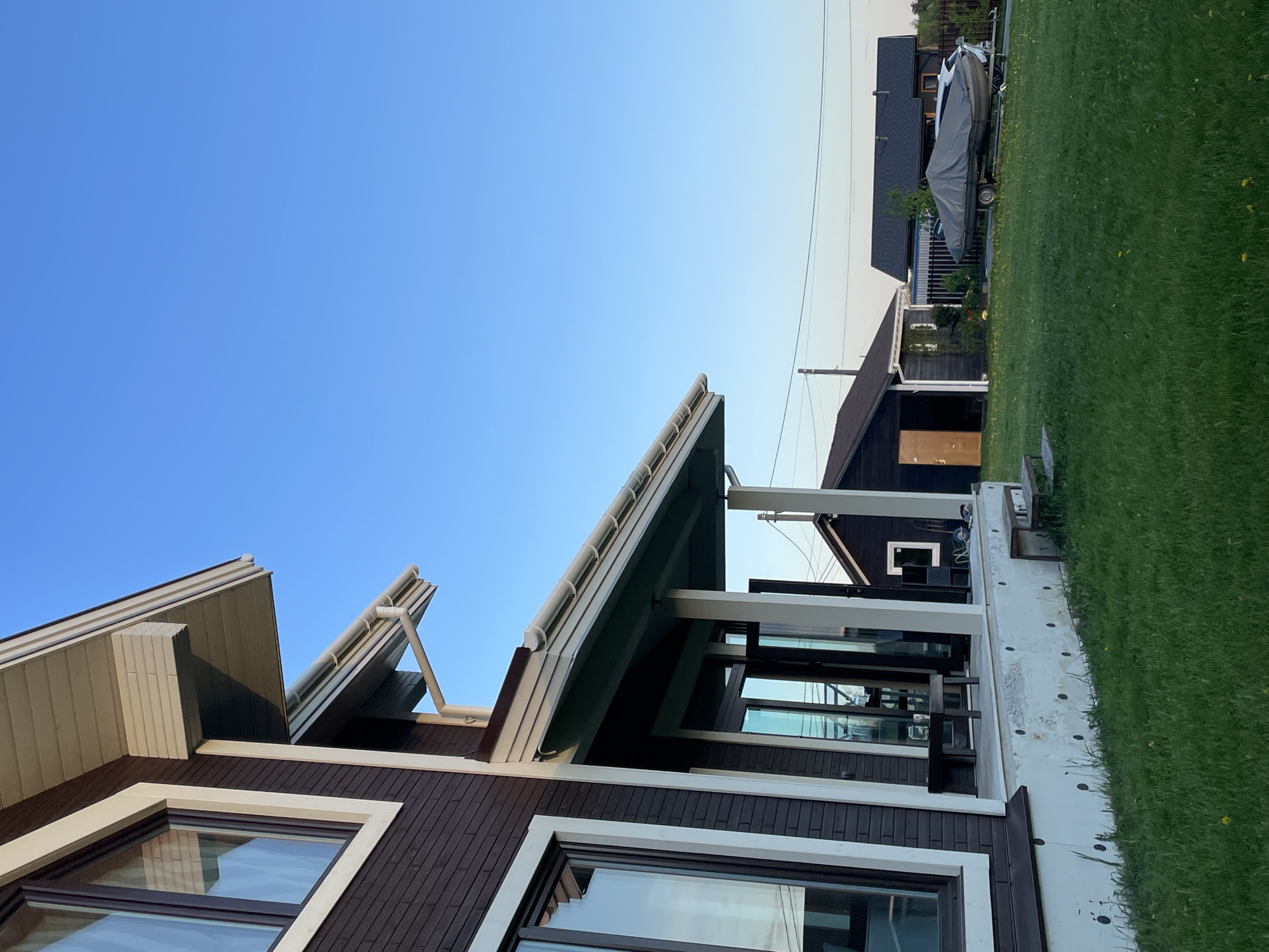 The image depicts a modern house in what appears to be a suburban or rural setting. The house has a dark brown exterior with white trim around the windows and roof. The roof is angular with multiple sections. Large windows reflect the surroundings. A covered patio area features black supports and possibly furniture visible through the windows. Adjacent to the house is a lawn and in the background, another building, potentially a garage or shed, and a boat on a trailer. The sky is a clear gradient of blue. The location is presumed to be Solov'evka, Russia, based on the prompt. There are no people visible in the image.