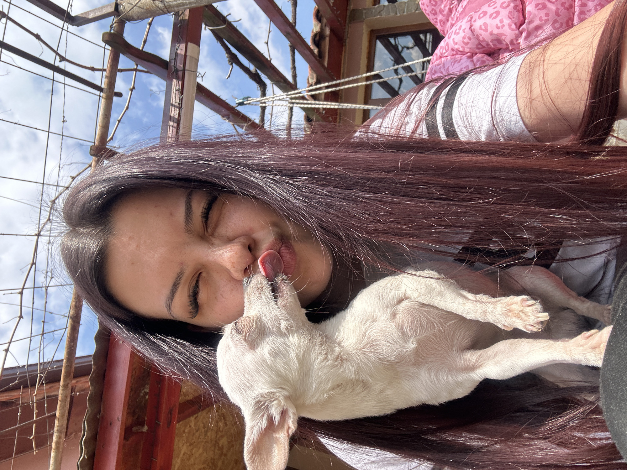 A woman with long, dark red hair and fair skin is holding a small, white dog. She is squinting and puckering her lips as the dog licks her face. The woman is wearing a white shirt with black stripes and a pink jacket. The scene is outdoors, evidenced by the presence of a metal trellis structure, a partially visible building, and a glimpse of a blue sky with clouds. Given the location provided (Chubra, Bulgaria), the building appears to be of a traditional architectural style.