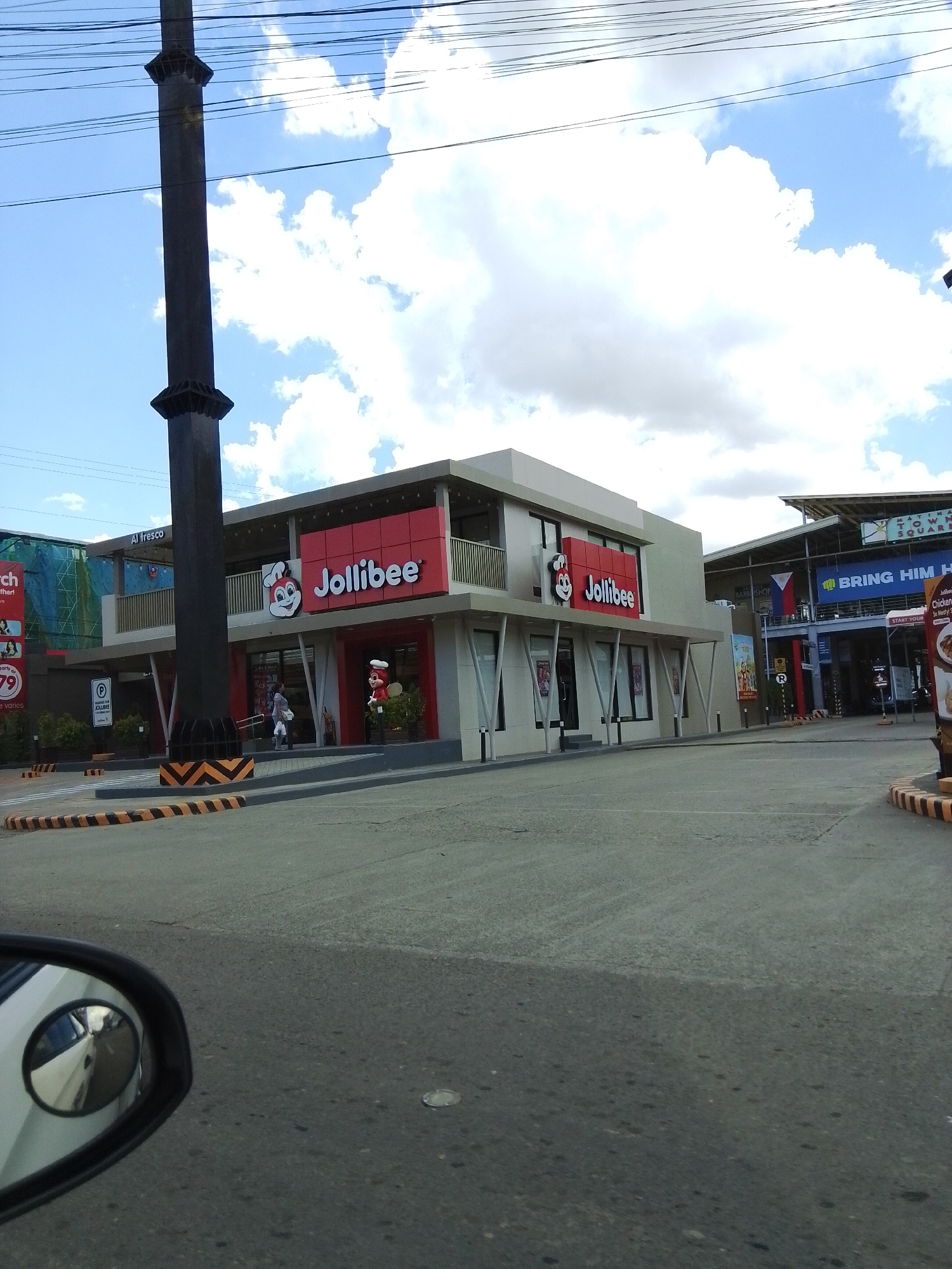 The image shows a Jollibee restaurant in Davao City, Philippines. The restaurant is a modern building with a red and white facade, and it features the Jollibee logo. There is a large, dark pole in the foreground, which appears to be a utility pole or part of a sign. The sky is blue with white clouds. In the background, there is a glimpse of a larger shopping complex with signage that includes "MATINA TOWN SQUARE." The image is taken from a low angle, possibly from a vehicle, as a car's side mirror is visible in the bottom left corner. There are a few people visible near the entrance of the Jollibee, and decorative elements like plants and a statue of a Jollibee character are also present. The area appears to be an outdoor public space with asphalt paving and yellow and black striped barriers.