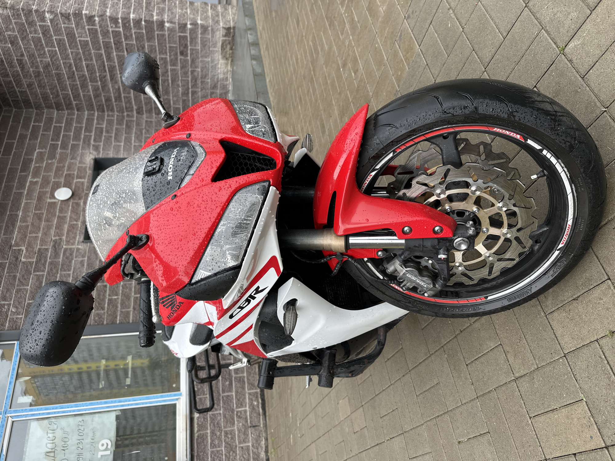 A red and white Honda CBR motorcycle is parked on a wet, paved walkway in front of a brick building. The motorcycle is a sportbike with a red fairing, white accents, and a black windshield covered in water droplets. The front wheel, with a silver brake disc, is prominent in the foreground, also showing water droplets. A black crash bar is attached to the motorcycle's frame. In the background, a window with signage is visible, and the brick wall of the building extends upwards. The overall scene suggests an outdoor setting on a damp day.
