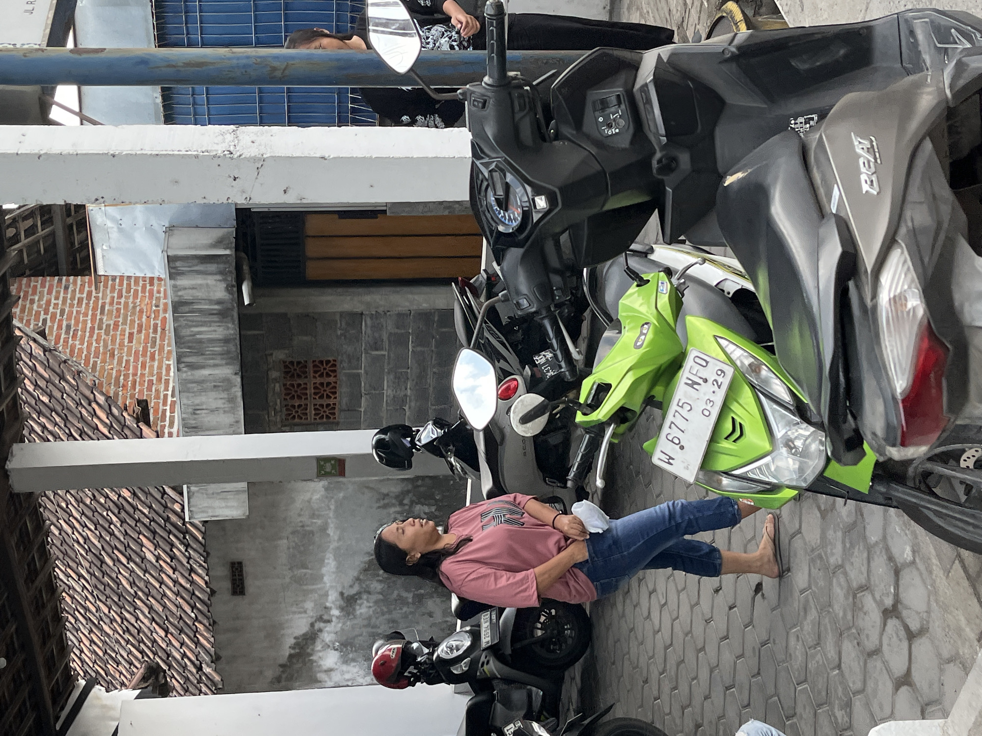 A woman in a pink t-shirt and blue jeans stands on a patterned tiled surface in front of several parked motorcycles. She is holding a white tissue in her right hand and has her left hand in her jeans pocket. One motorcycle in the foreground is bright green, with a license plate reading "W 6775 NFQ 03.29". Behind her, other motorcycles, some with helmets on them, are visible. A brick building with a tiled roof is in the background, with a white pillar to the right of the woman. The scene appears to be an outdoor area, possibly a parking lot, in Kepatihan, Indonesia, as suggested by the license plate.