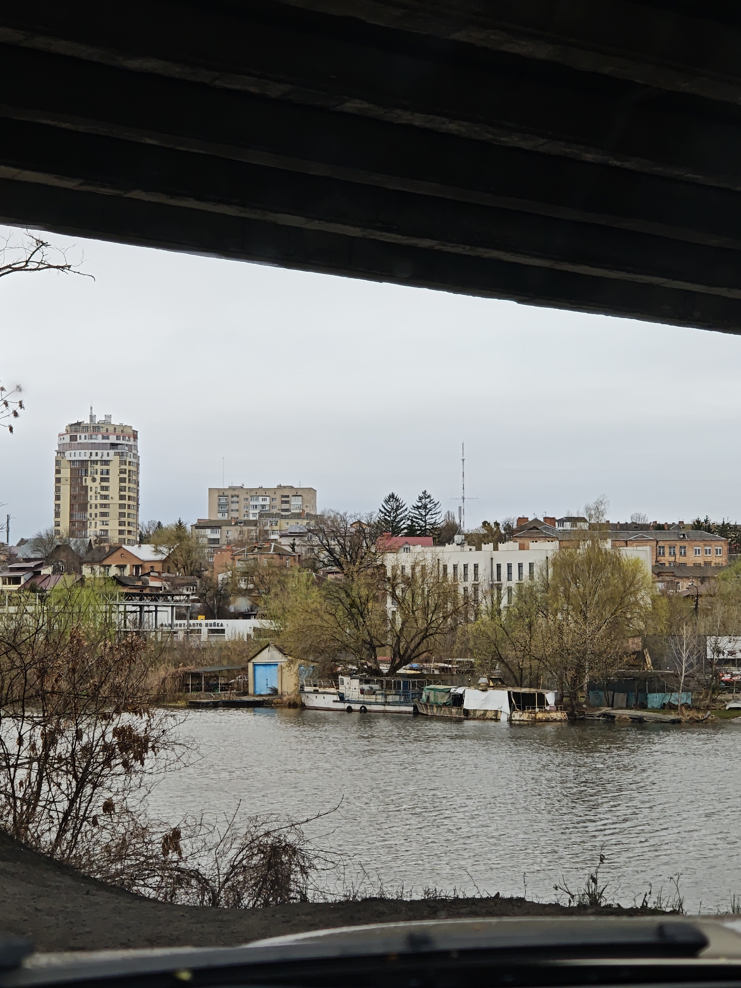 View of the city of Vinnytsia, Ukraine, from underneath a bridge. In the distance, tall apartment buildings are visible on a hill. Closer to the viewer, a river or body of water is seen with several small boats docked along the shore. The foreground includes bare trees and branches, indicating it may be late autumn or early spring. The sky is overcast, suggesting a cool or rainy day. The image is likely taken from inside a vehicle.