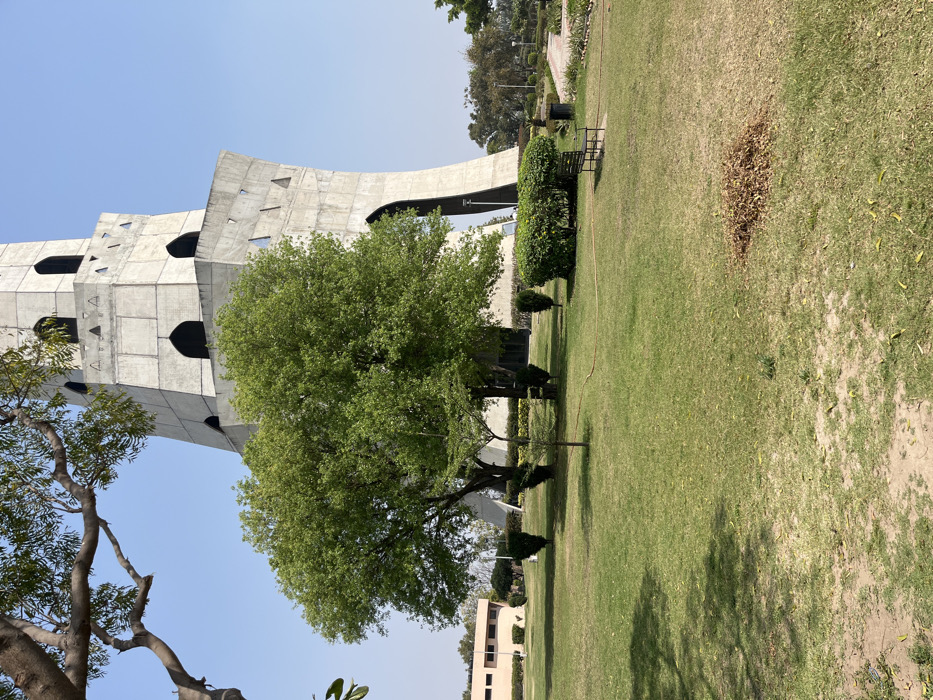 The image shows a tall, concrete tower with a unique architectural style in Sahibzada Ajit Singh Nagar, India, also known as Fatehgarh Sahib. The tower is situated in a grassy park. The structure is made of concrete with a design featuring rectangular windows. There are two large trees near the base of the tower. A small bench is visible in the foreground, sitting on a grassy area. The lawn appears well-maintained with green grass, while the surrounding ground is covered in brown leaves and dirt patches. A small bush sits in front of the bench. There is a large tree in the background to the left of the tower and several other trees at the edge of the image, indicating the park is fairly large. The image is taken from a low angle, looking up at the tower. The weather is sunny and clear, with a blue sky visible in the background.
