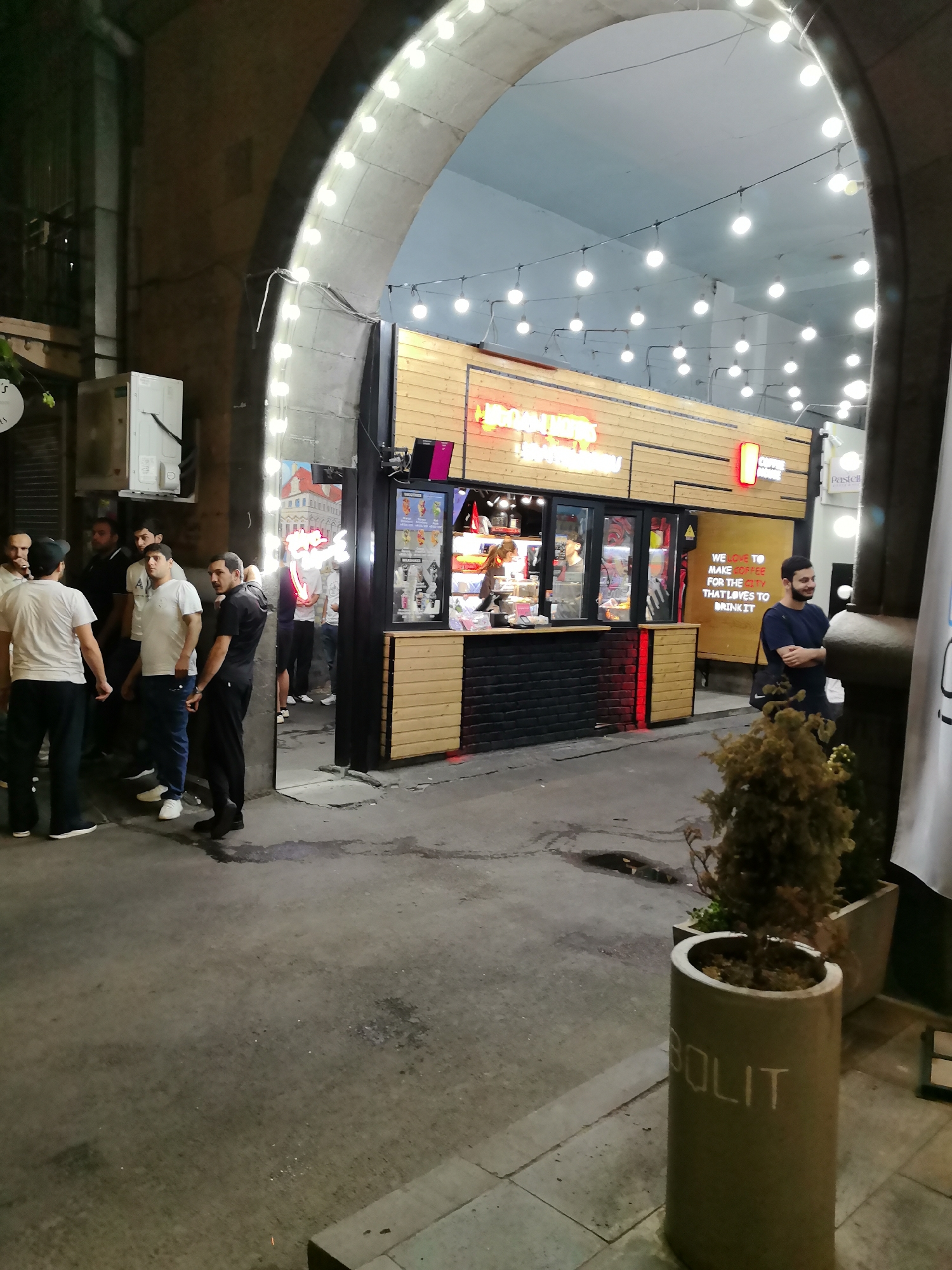 A group of men stand outside a cafe located under an arched walkway in Yerevan, Armenia. The cafe has a wooden facade with a black brick base and red neon signage. String lights are strung across the ceiling of the walkway, illuminating the scene. The ground is paved with concrete and some areas appear wet. A cylindrical planter with a small tree is positioned in the foreground, with the word "SOLIT" stenciled on its side. Several men are gathered near the entrance of the cafe, some appearing to be customers or employees. One man in a dark blue t-shirt stands with his arms crossed, looking to the right. Another man in a black shirt and pants is looking towards the viewer. The overall atmosphere suggests a casual evening gathering in a pedestrian area.