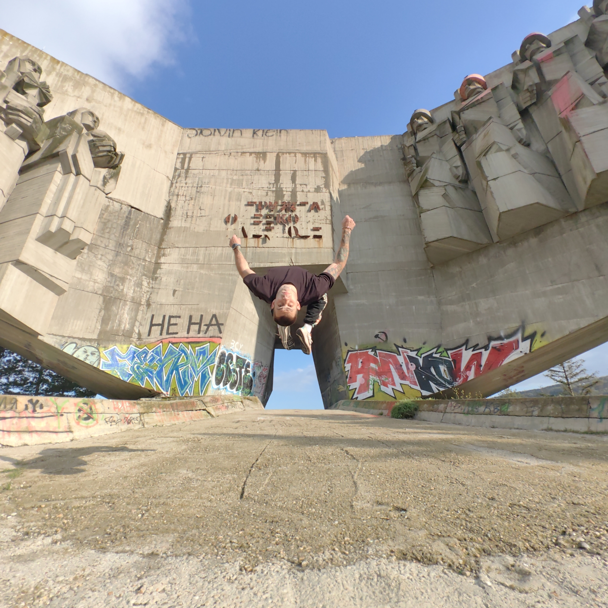 The image displays a man captured in mid-air, performing an acrobatic flip or jump, centrally framed within a large concrete monument in Varna, Bulgaria. He is wearing a dark t-shirt, dark pants, and light-colored sneakers, with visible tattoos on his arms, and looks directly at the camera with a focused expression, fists clenched.
The monument is a massive, brutalist concrete structure featuring abstract, blocky sculptural figures on its left and right sides, some with faded red accents on their headwear. The architecture is imposing and weathered, consistent with Soviet-era design. The ground leading up to the monument consists of a gravelly, dirt path. Graffiti extensively covers the lower sections of the concrete walls and the ground-level barriers, adding a contemporary, urban layer to the historic site.
The scene takes place on a bright, sunny day with a clear blue sky and minimal cloud cover. Through the opening in the monument, where the man is positioned, additional blue sky is visible. Green vegetation, including bushes and distant trees, can be seen at the base of the structure and on the surrounding hills.
Visible text includes faint black graffiti spelling "DOLVIN KLEIN" (possibly a variation of "Calvin Klein") and fragmented reddish-brown characters that resemble "PWOK-A ZBKO" on the upper central wall. Below that, "HE HA" is written in black on the lower left wall, amidst various other colorful and illegible graffiti tags.