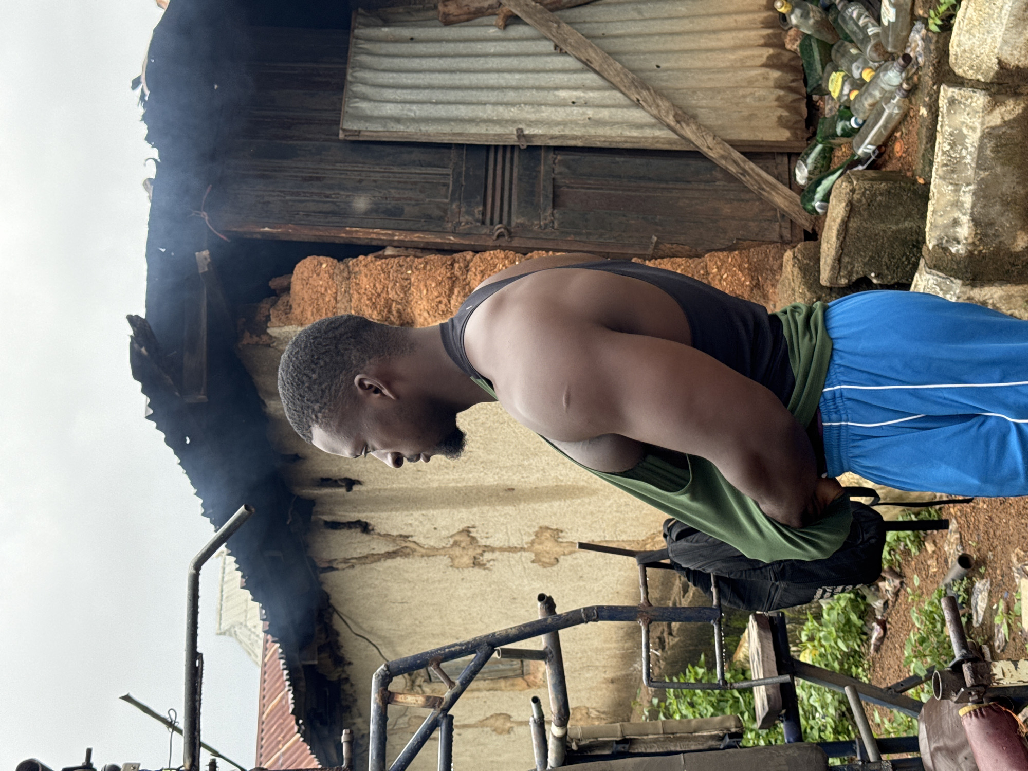 In Akwanga, Nigeria, a muscular Black man stands outdoors next to a dilapidated structure and gym equipment. He is wearing a black tank top and blue athletic shorts. His right arm is bent, and he appears to be adjusting his clothing, with a green tank top draped around his waist. Behind him, the building's roof is partially collapsed and smoking, suggesting a recent fire. Discarded glass bottles are scattered on the ground near the building's entrance, which is made of corrugated metal. The environment appears to be rural or impoverished, with overgrown vegetation and a general sense of disrepair. The sky is overcast.