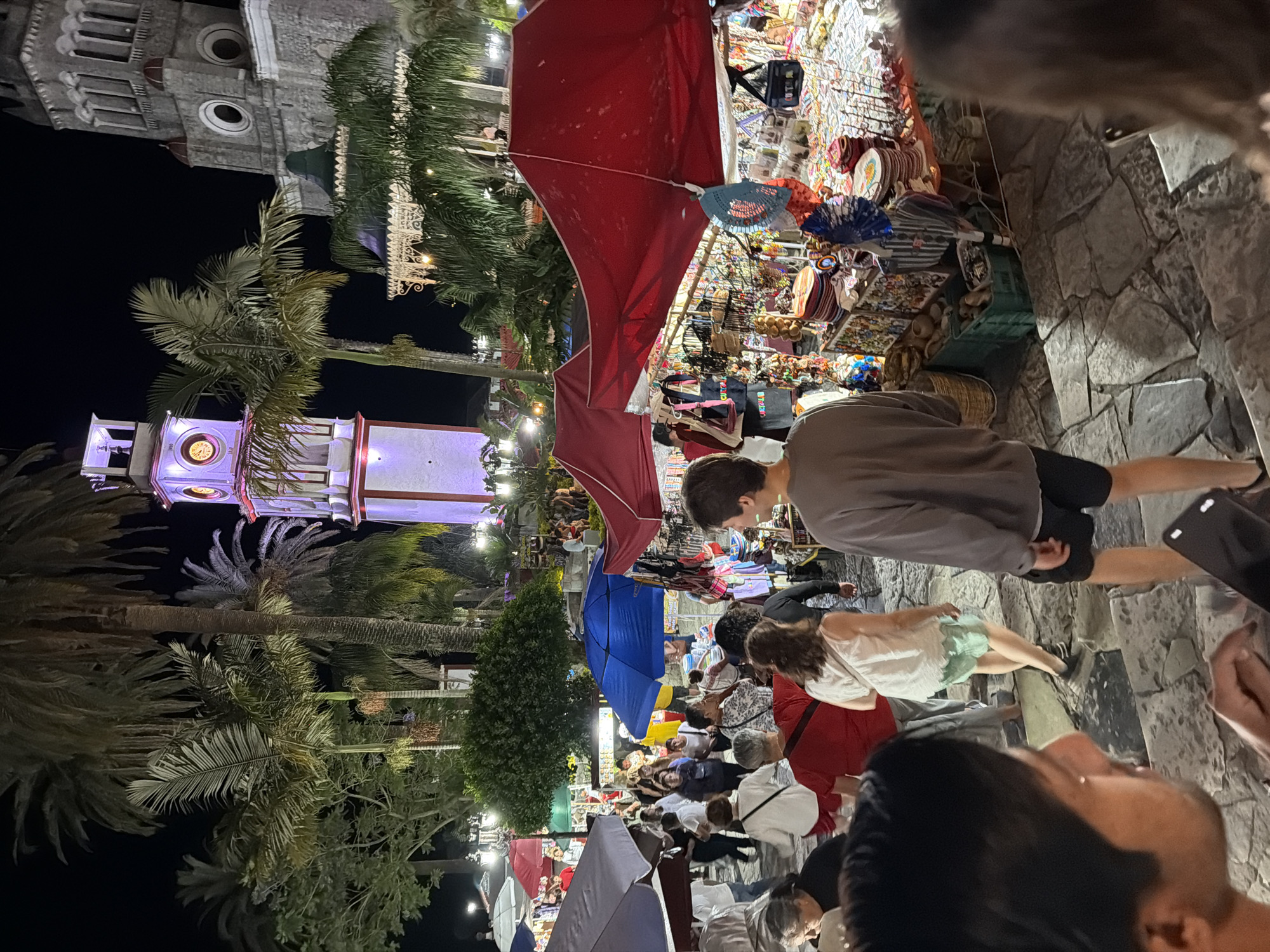 The image is an eye-level, nighttime photograph of a market in Cuetzalan, Mexico. A large crowd of people are walking through the market, looking at goods displayed on tables and under awnings. Merchandise for sale includes jewelry, clothing, and souvenirs. A tower with a clock is visible in the background, illuminated with colored lights. Palm trees are also visible in the background, and the sky is dark. The ground is made of cobblestones.