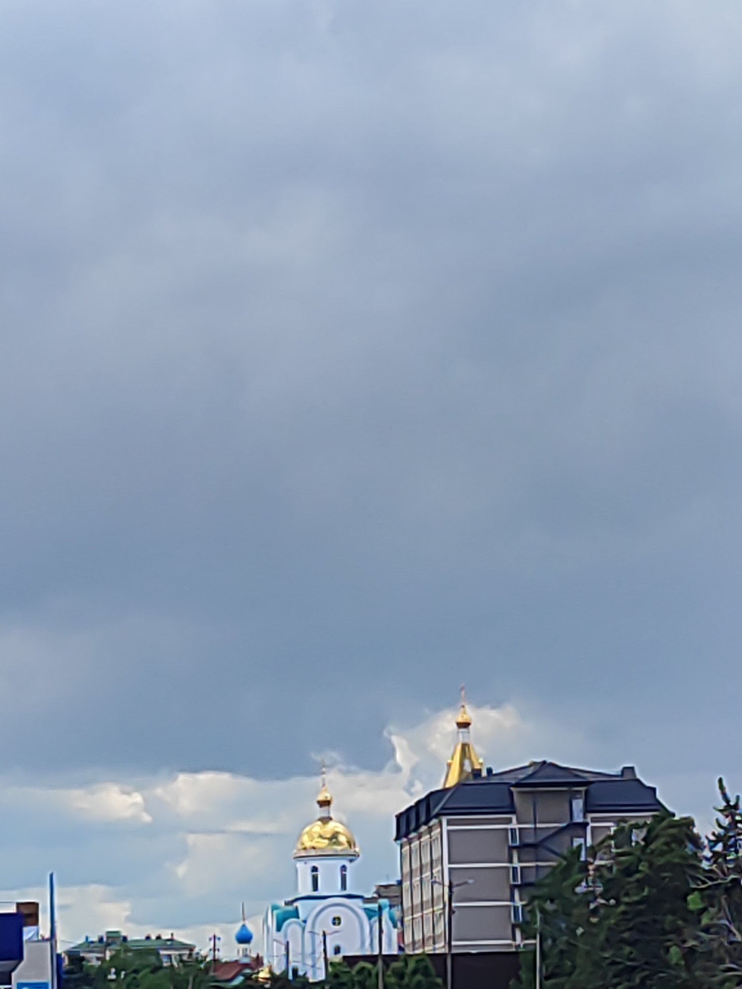 The image depicts a cloudy sky over Krasnodar, Russia. In the lower portion of the frame, a cityscape is visible, including a church with golden domes and a modern building. Vegetation appears in the foreground.