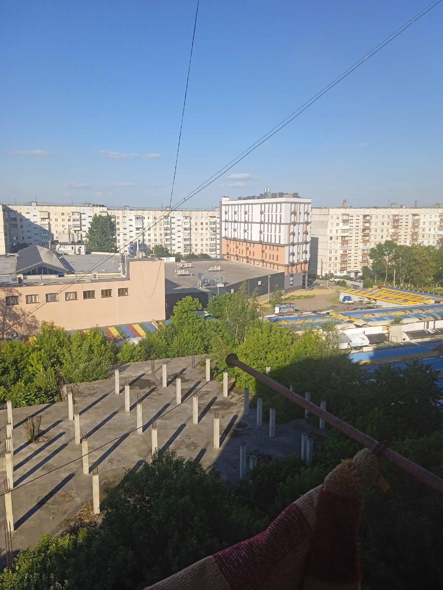 The image depicts a view of an urban environment in Chernivtsi, Ukraine, likely from an elevated vantage point. In the foreground, there is a concrete structure with vertical pillars and shadows cast by the sun, suggesting it might be a foundation or an unfinished building. Dense green foliage is visible below and around this structure.
In the midground, several buildings are visible. One prominent building is a modern, multi-story structure with a white facade and some orange accents. Adjacent to it are several older, light-colored apartment buildings with numerous windows. A peach-colored building with rectangular windows is also present, featuring a rainbow-colored awning or roof structure underneath it. Various air conditioning units are mounted on the exteriors of some buildings.
The background is dominated by a clear blue sky with a few wispy clouds. Power lines are strung across the sky, and a few small birds appear to be perched on them. Beyond the apartment buildings, more residential structures extend into the distance. The scene suggests a typical daytime view of a residential and commercial area in a Ukrainian city. There are no people visible in the image, nor any specific activities taking place. The lighting indicates it is likely morning or late afternoon due to the long shadows.