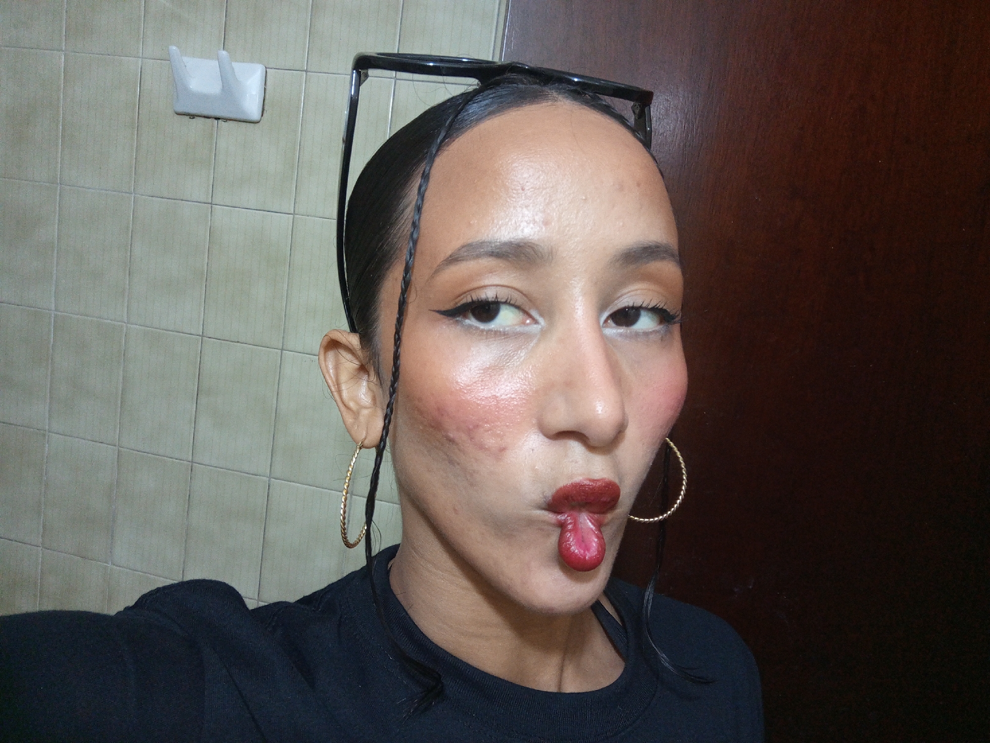 A woman with dark hair pulled back and adorned with braids is taking a selfie. Her hair is slicked back, and a pair of glasses is perched on top of her head. She is wearing gold hoop earrings, black eyeliner, blush, and dark red lipstick. Her tongue is sticking out, pursed into a playful expression. She is wearing a black shirt. The background includes a tiled wall on the left side with a white plastic towel rack and a dark wooden surface on the right side. The location, Porlamar, Venezuela, cannot be confirmed from the image alone.