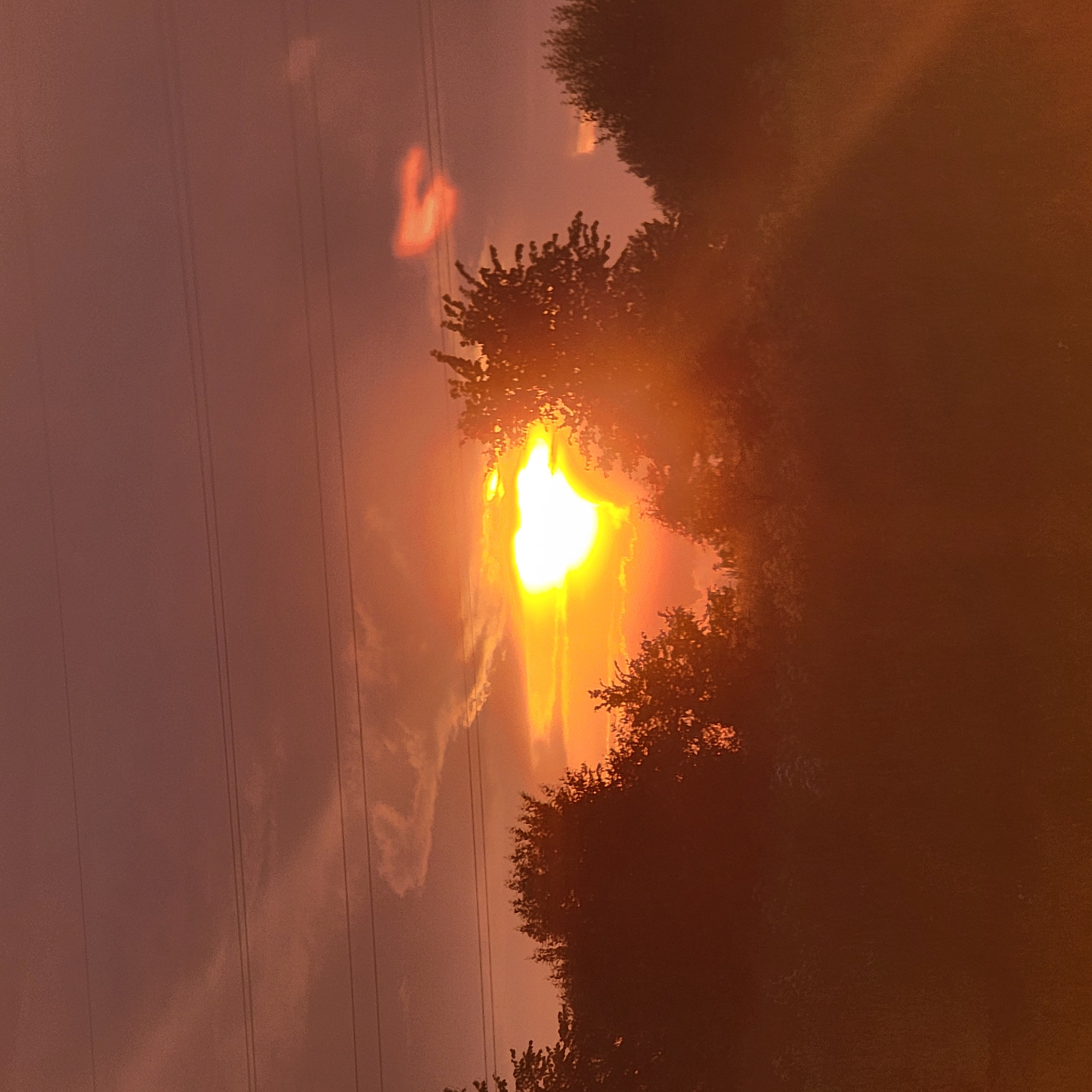 This photograph captures a dramatic sunset or sunrise, likely a solar eclipse, viewed through silhouetted trees. The sun, partially obscured by what appears to be the moon, glows with an intense, fiery orange and yellow light. Wisps of clouds are illuminated by the sun's brilliance, creating ethereal streaks against the dusky sky.
The foreground is dominated by the dark, textured shapes of trees, their branches reaching upwards and framing the celestial event. In the background, thin, dark lines suggest the presence of power lines, hinting at an outdoor, possibly rural or suburban, setting. The sky transitions from a muted reddish-brown near the horizon to a lighter, almost pinkish hue higher up.
There are notable light artifacts in the image, such as bright, amorphous shapes in the sky, which could be lens flares or reflections. The overall mood is one of awe and wonder, amplified by the unusual spectacle of the sun being partially hidden. The low angle of the light and the rich colors strongly indicate either early morning or late evening. The provided location context, Ufa, Russia, places this event geographically. No text is visible in the image.