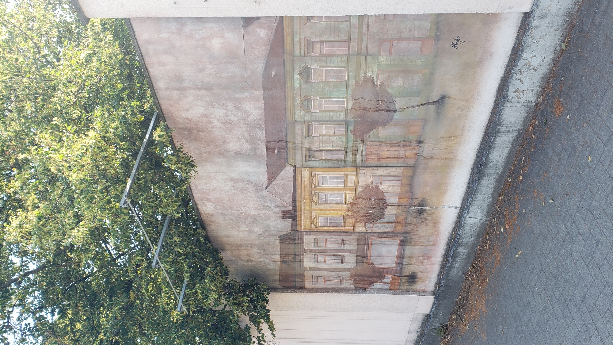 A mural depicts a row of old European-style buildings with arched windows and colorful facades, rendered in a faded, somewhat impressionistic style. The buildings are reflected in a watery surface below, suggesting a flooded street or a rainy day. Small, sparse trees with bare branches are painted in front of the buildings. The mural is painted on a concrete wall, and a section of a white building is visible to the left, and a white pillar is on the right. Above the mural, a large tree with lush green leaves overhangs the wall, with metal structures visible through the branches, possibly part of a trellis or support system. The ground in front of the wall is paved with grey interlocking bricks, and scattered fallen leaves are visible on the paving stones and at the base of the wall. The mural is signed with the name "Hovka" and the year "18". This mural is located on Masarykova Street in Prešov, Slovakia.