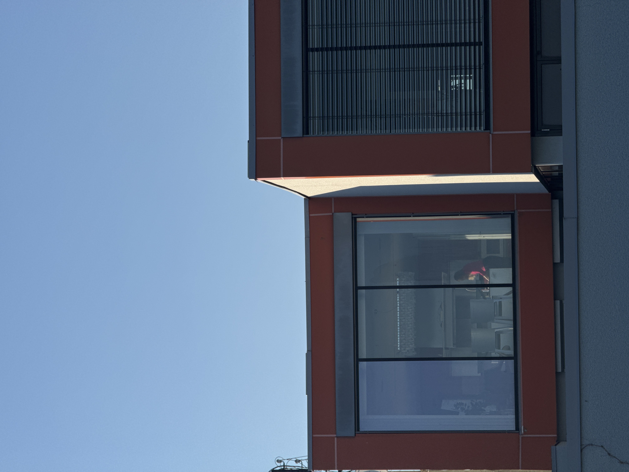 The image shows a building with a distinct architectural style. The building features a combination of orange-red panels with grey accents and large windows. One window shows a person wearing a red shirt. The building’s design incorporates geometric shapes and straight lines. The surrounding environment includes a clear blue sky. The location appears to be an urban area.