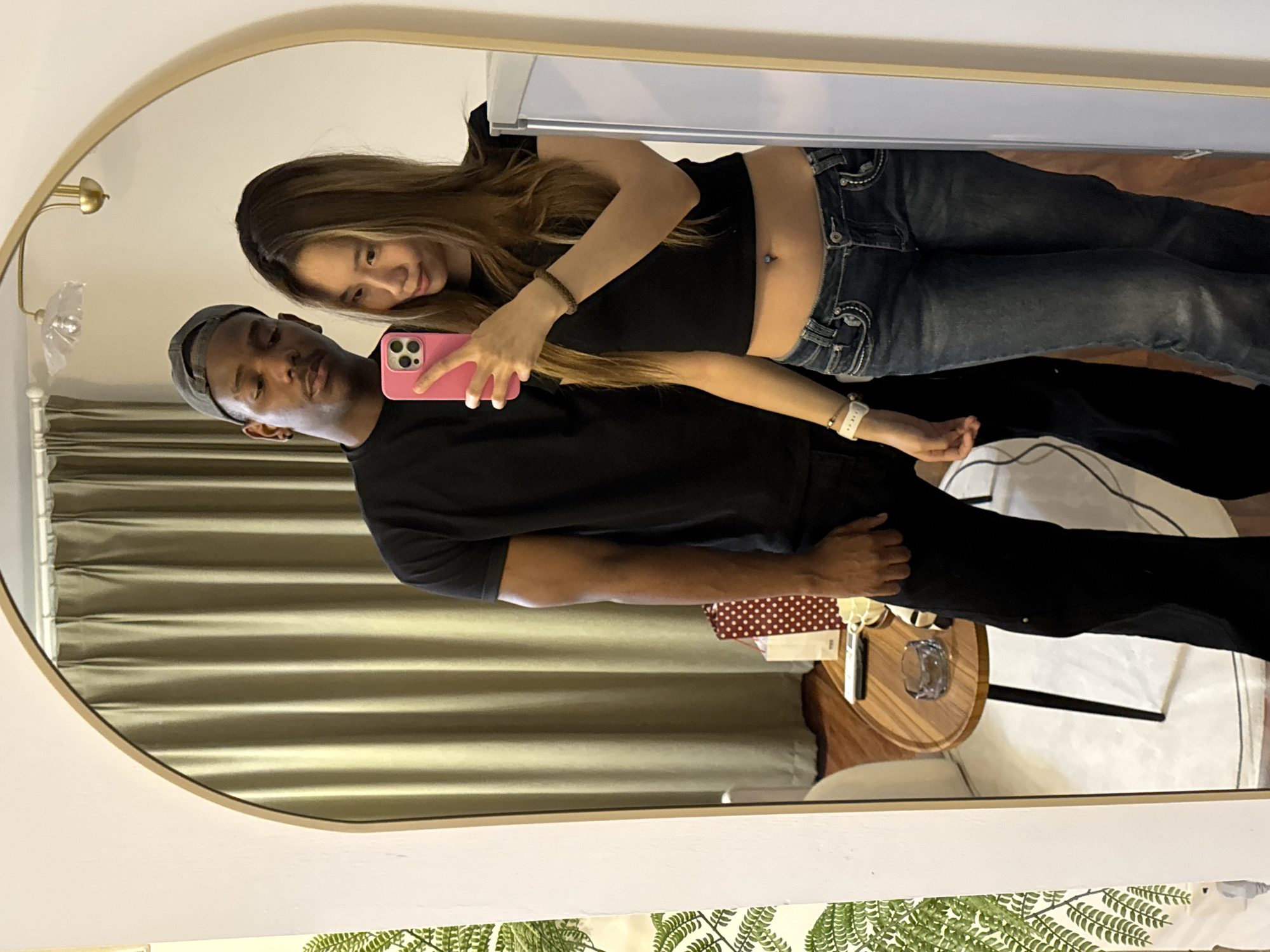 The image shows a selfie taken in a mirror. A man and a woman are visible. The man is wearing a dark shirt and a cap. The woman is wearing a dark top and jeans. She is holding a pink smartphone, presumably taking the picture. The background shows curtains, a side table with a glass and other items, and what appears to be a bed. It is difficult to determine the specific location in Yuzhong District, China from the image.