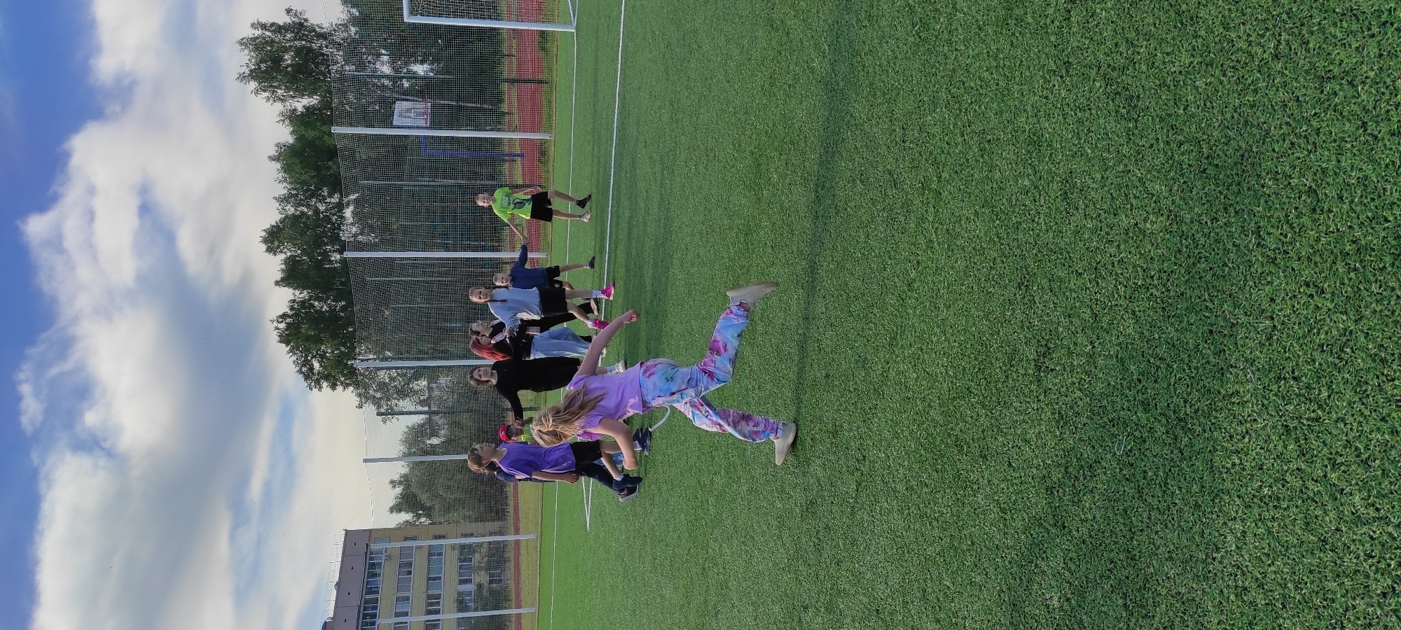 A group of children are playing on a green artificial turf field, likely a soccer field, in Noviy, Russia. The sky is cloudy with patches of blue. In the background, a multi-story building and trees are visible. Some of the children are in motion, suggesting active play or a game. One child in the foreground is running, her back to the camera, wearing colorful patterned pants and a light purple t-shirt. Other children, boys and girls, are grouped together, some appearing to be holding hands or interacting. Sports equipment, such as a soccer goal, is present on the field. The scene depicts a casual outdoor activity.