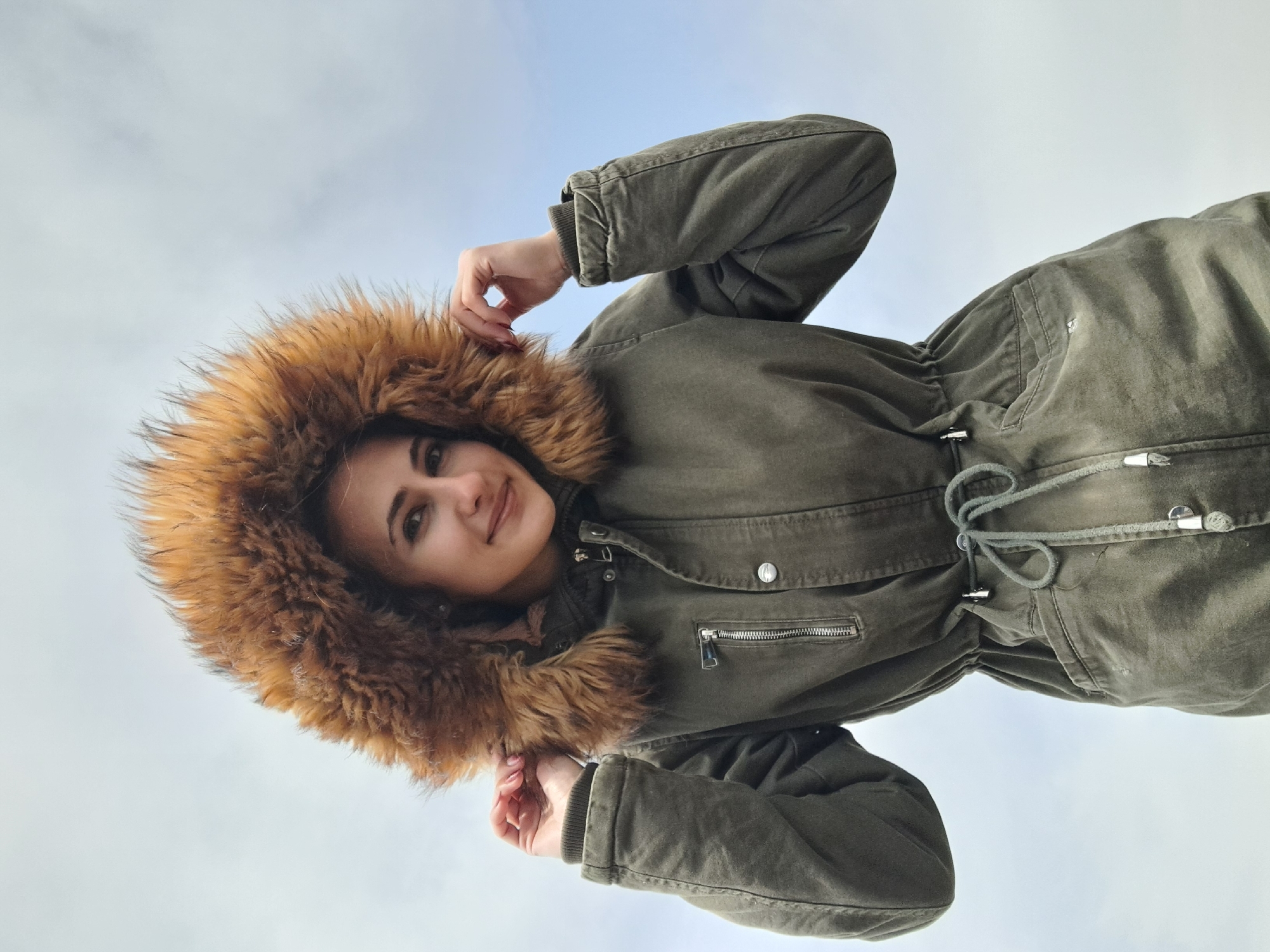 A young woman with brown hair and fair skin is wearing a green parka with a large fur-trimmed hood. She is holding the fur of the hood with both hands. The background is a cloudy sky. The location is not discernible from the image.