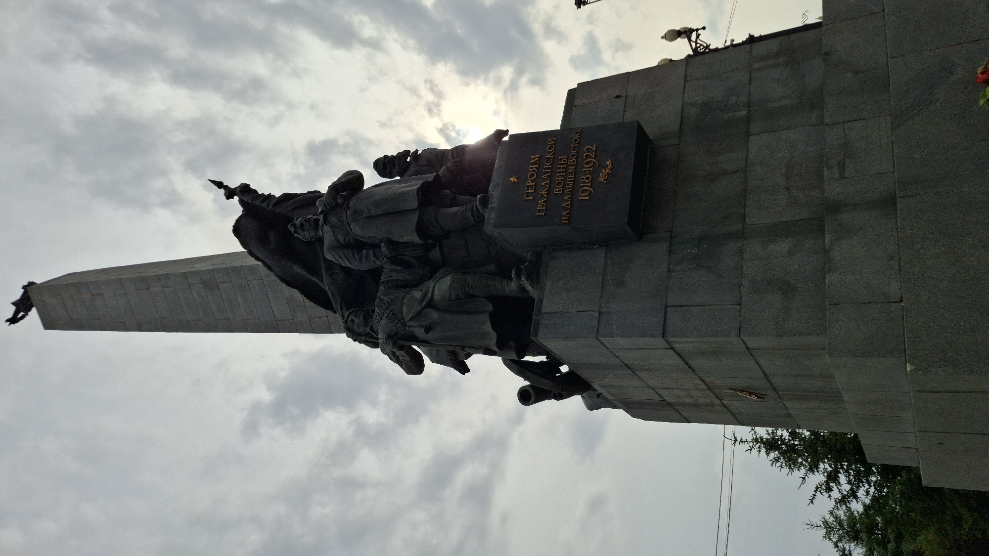 The image shows a low-angle view of the monument to the heroes of the Civil War in the city of Khabarovsk, Russia. The monument is made of granite and features several bronze sculptures of soldiers and a tall obelisk topped with a figure. A plaque on the monument reads "To the heroes of the Civil War in the Far East 1918-1922". The sky is overcast with clouds, and the sun is partially visible, creating a bright glare. Trees and streetlights are visible in the background.