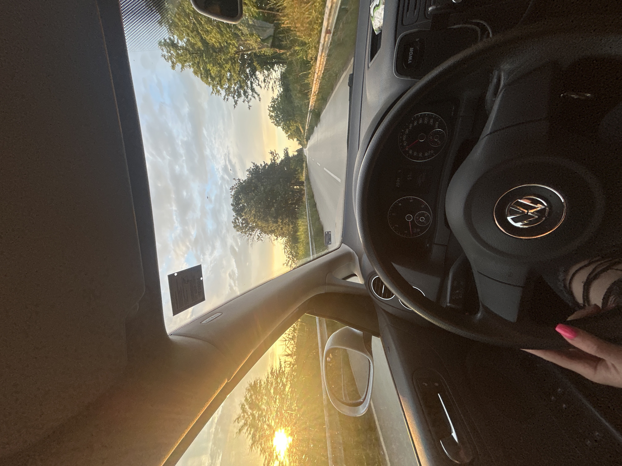 The image depicts an internal view from a moving vehicle on a road in Milkovitsa, Bulgaria, during sunset. The time displayed on the vehicle's dashboard is 20:34, with another indicator showing "51". A hand with hot pink nail polish is visible in the lower-left, gripping the steering wheel, which features a chrome Volkswagen emblem. Part of a leg, clothed in ripped black jeans, is also visible. The driver's side door panel, equipped with a chrome handle and control buttons, is prominent. An A-pillar with an "AIRBAG" label and the exterior side mirror reflecting the road are on the left. A sticker is affixed to the upper right section of the windshield. A device labeled "YWOR" is located on the dashboard, beneath a white plastic item.
Externally, a paved two-lane road with distinct white lane markings extends into the distance. Metallic guardrails line both sides of the road, bordered by dense green trees and tall grass. The sun is low on the left horizon, creating a significant glare and bathing the environment in a golden light. The sky above is partially cloudy, revealing patches of blue. Small, unidentifiable airborne objects are visible against the sky. No other vehicles are immediately discernible on the road.