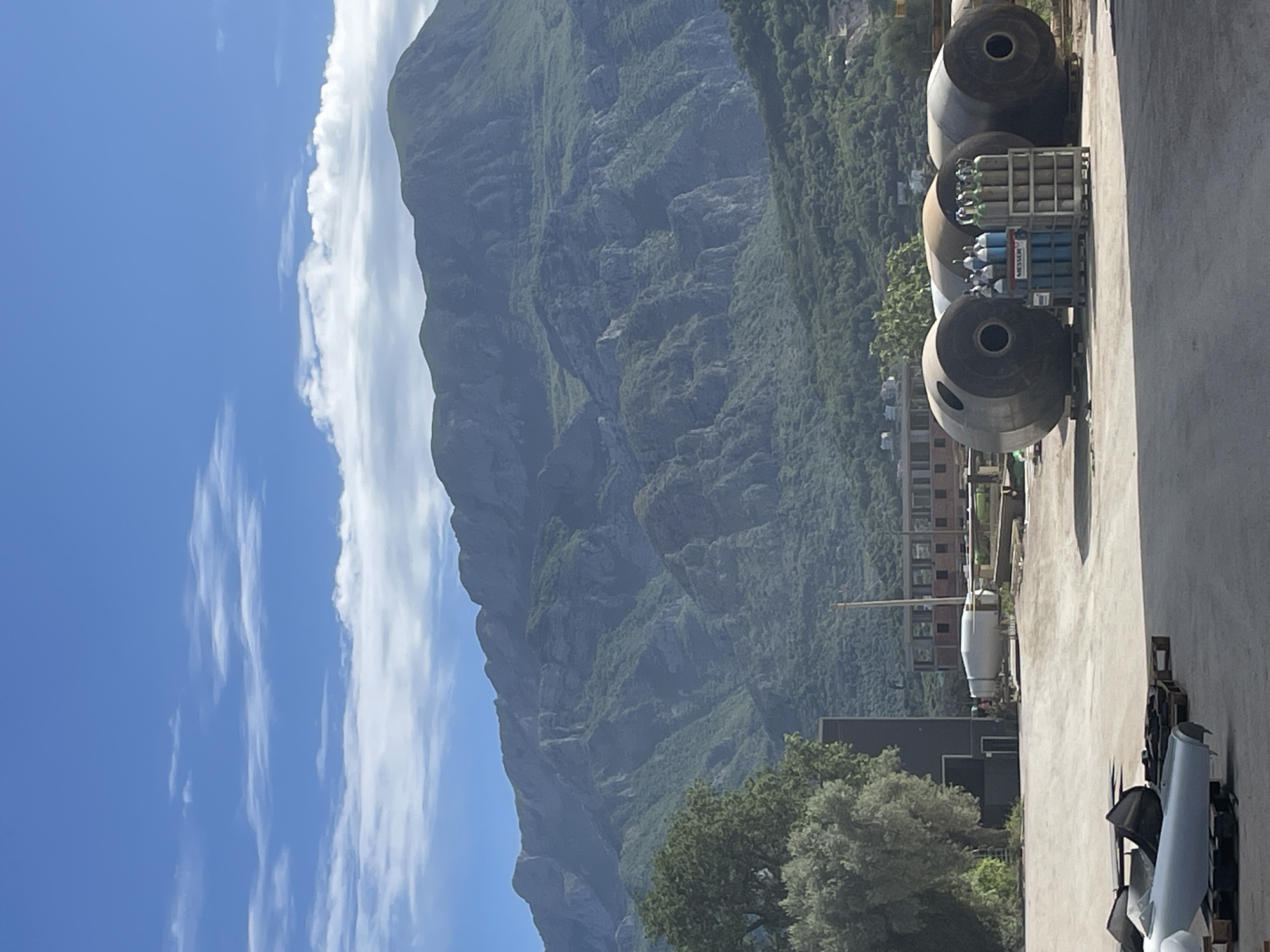 The image shows a view in Čeluga, Montenegro, with a mountain dominating the background under a blue sky with clouds. In the foreground, on what appears to be a concrete surface, are large, dark cylindrical objects with open ends, possibly industrial or construction materials. A cart with numerous smaller gas cylinders is positioned next to these cylindrical objects. Further along the concrete surface, there is a lighter colored tank-like object and other scattered pieces of equipment or materials. Buildings can be seen amidst the greenery in the middle ground. The overall scene suggests an industrial or construction-related area with the backdrop of a mountain landscape.