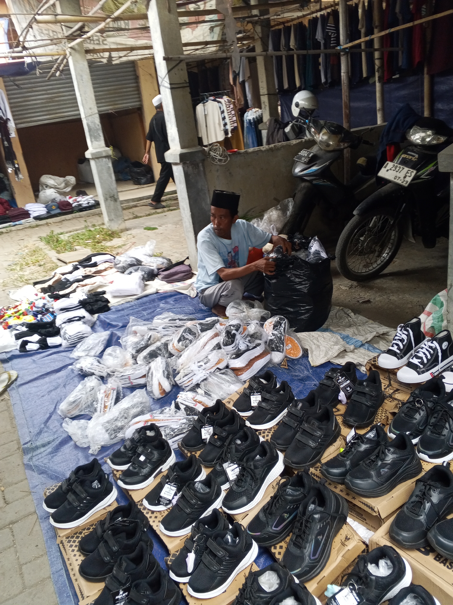 A street market scene unfolds in Serang, Indonesia. The foreground is dominated by a large blue tarp spread on the ground, displaying a variety of footwear. Numerous pairs of black athletic shoes are neatly arranged on cardboard boxes and directly on the tarp, some still in plastic packaging. Among these are also white and black sneakers, some with laces, others with velcro straps. To the left, a pile of folded clothing items in white and black is visible, along with some colorful items on a smaller piece of fabric.
In the mid-ground, a man wearing a light blue t-shirt and dark pants is seated on the tarp, sorting through a large black plastic bag. His attention is focused on the bag's contents, suggesting he is preparing or organizing merchandise. Behind him, two motorcycles are parked. One is dark, and the other appears to be a lighter color with a license plate visible.
Further back, a structure with a corrugated metal roof provides shade. Clothing items hang from a rack in the background, and a person wearing a white skullcap and black clothing walks away from the camera, towards the left side of the frame. The overall environment suggests an open-air market, likely a temporary setup for selling goods. The presence of motorcycles indicates this is an accessible area, possibly a roadside or a dedicated market space.