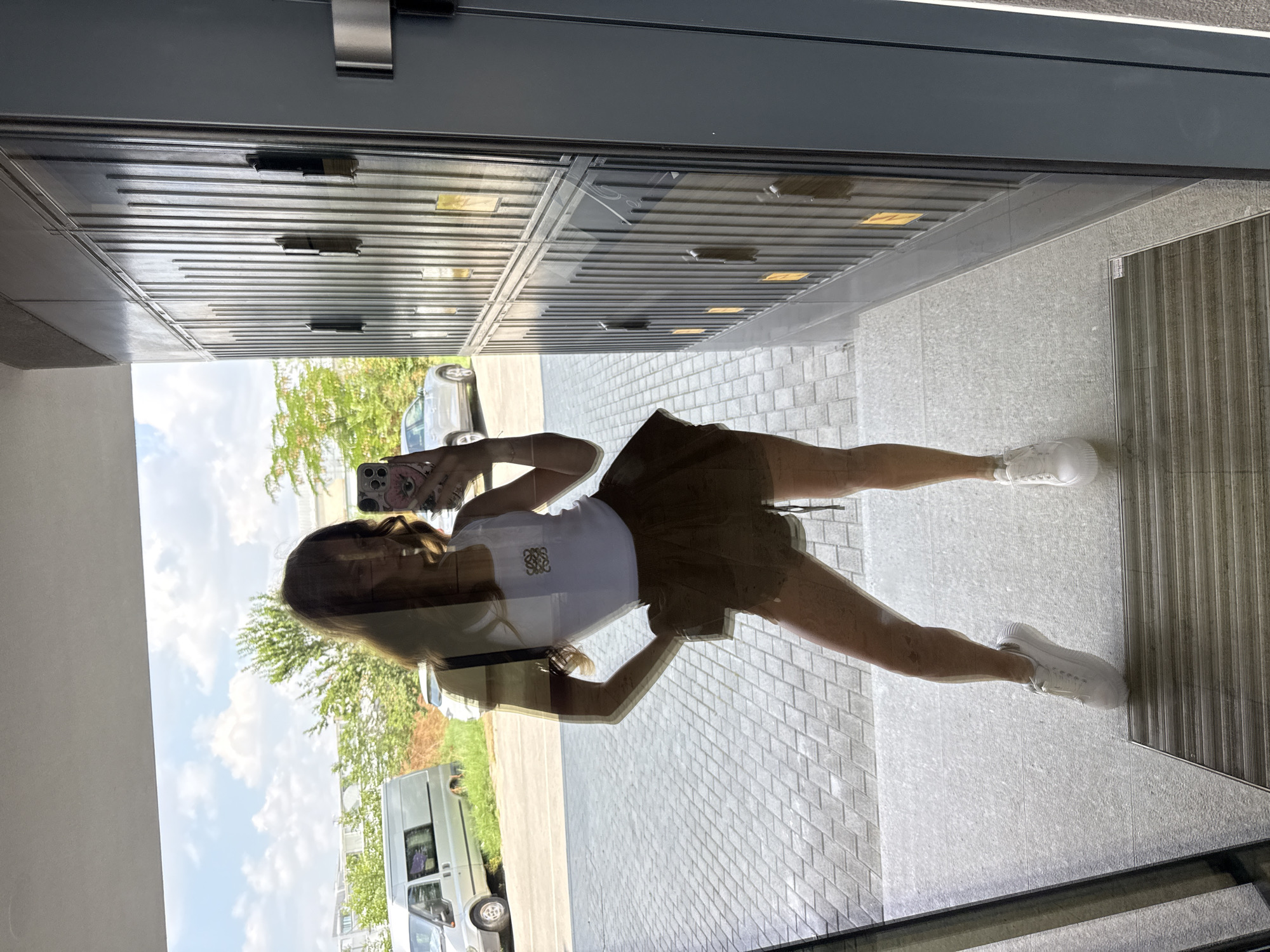 In Plovdiv, Bulgaria, a young woman, seen in a mirror reflection, stands on a tiled walkway. She is wearing a white tank top with a small logo, a brown ruffled skirt, and white sneakers. She is holding a smartphone, capturing her own image. Behind her, a reflection shows a street with parked cars and trees under a cloudy sky. To the right of the woman in the reflection, a grey wall with vertical grooved panels and multiple small doors is visible. The image appears to be taken in an outdoor, urban setting.
