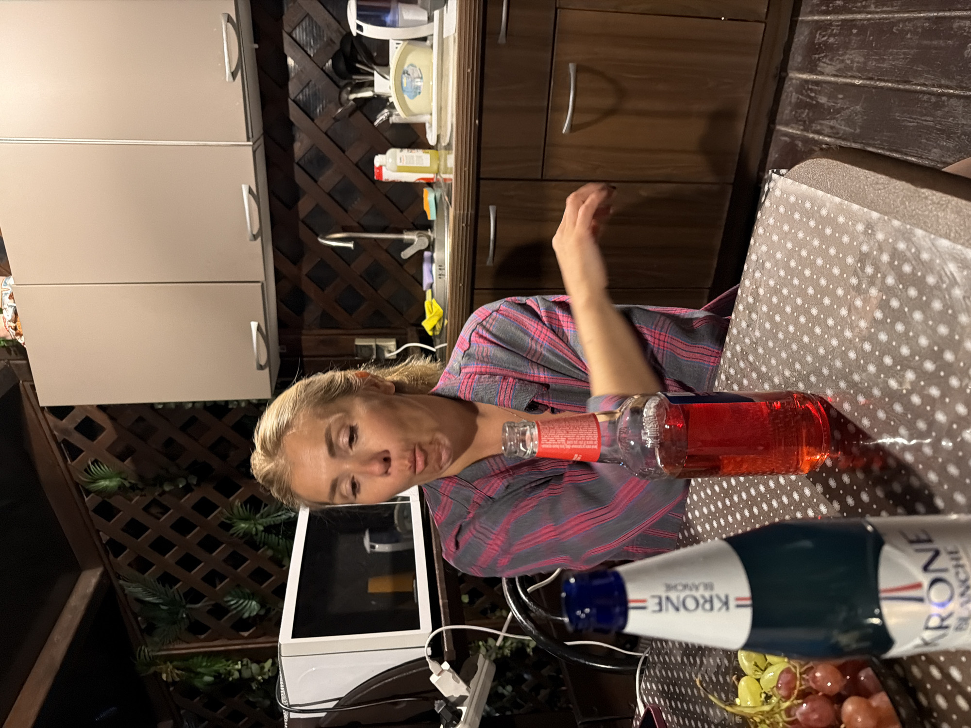 A Caucasian woman with blonde hair pulled back is sitting at a table in what appears to be an outdoor kitchen or patio area. She is wearing a pink and gray plaid shirt and has a surprised or amused expression, with her mouth open. In front of her on the table are two bottles of drink and a bowl of grapes. One bottle is dark green and white and labeled "KRONE BLANCHE." The other bottle is clear glass and contains a red liquid, with a label that is partially visible. The background shows wooden lattice work and kitchen cabinets. The scene is set in Sochi, Russia.