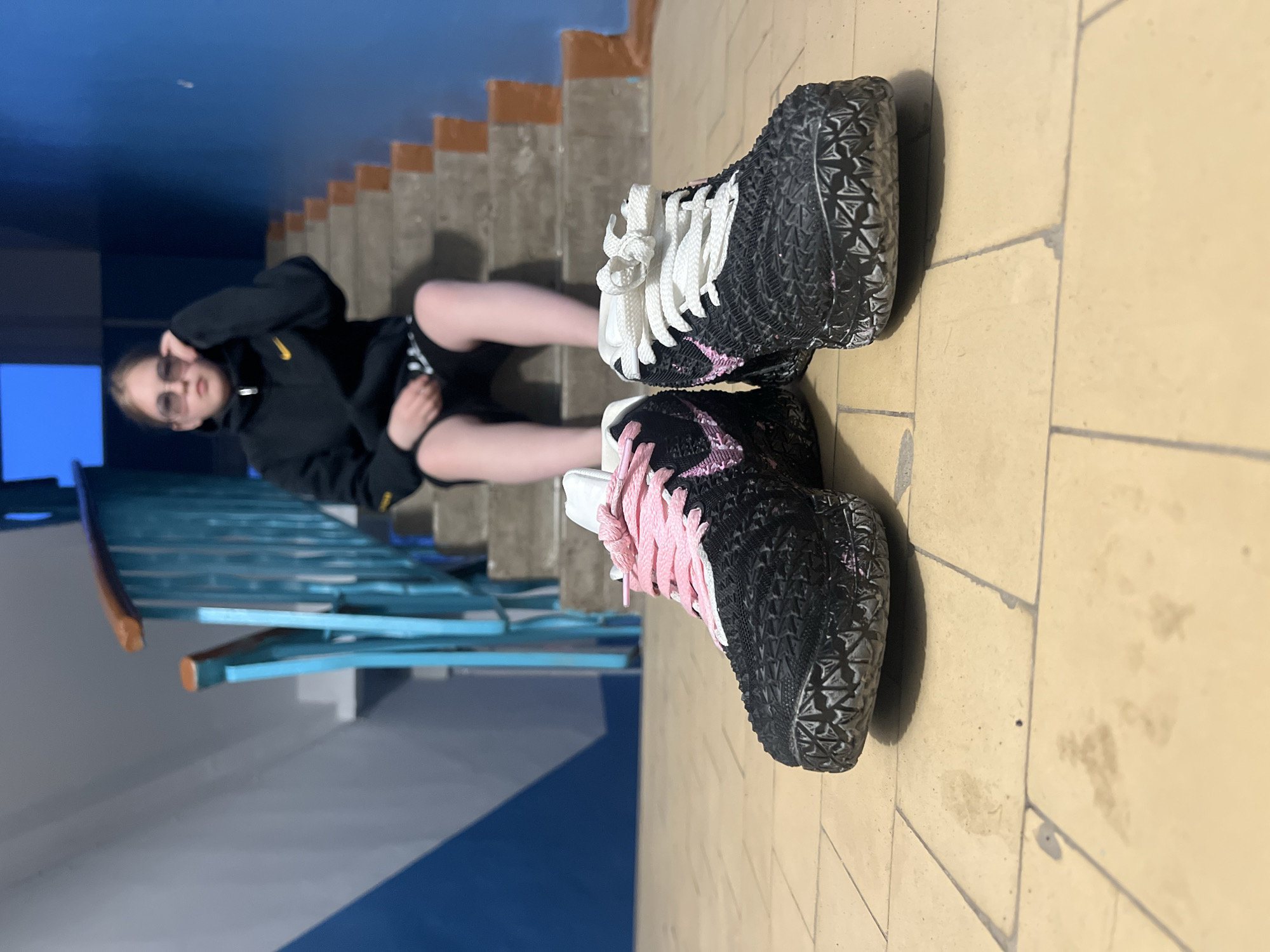 An indoor scene in Chaikovsky, Russia, features a young person reclining on a staircase, with two pairs of sneakers prominently displayed on the tiled floor beside them. The image is captured with artificial lighting, casting visible shadows.
The central subject is a young individual, likely female, wearing a black hoodie with a yellow Nike swoosh logo on the chest, black shorts, and light-colored tights or leggings. They wear dark sunglasses, obscuring their eyes, and have light-colored hair. Their left hand is raised towards their face, possibly adjusting the sunglasses or posing. They are positioned partially on a concrete staircase and partially on a landing, with their legs extended.
Adjacent to the person, on a beige tiled floor, are two black sneakers. Both shoes feature a textured, possibly knitted or molded, black upper and a thick, patterned sole. One sneaker has white laces and white detailing, while the other is equipped with bright pink laces and pink accents. They are arranged side-by-side, appearing to be intentionally displayed.
The background consists of a concrete staircase with light brown or orange risers, a distinctive light blue metal handrail with horizontal bars, and blue-painted walls. The overall setting suggests a public or institutional building interior, such as a school or community center. The lighting is somewhat dim, highlighting the subjects against the blue and beige surroundings.