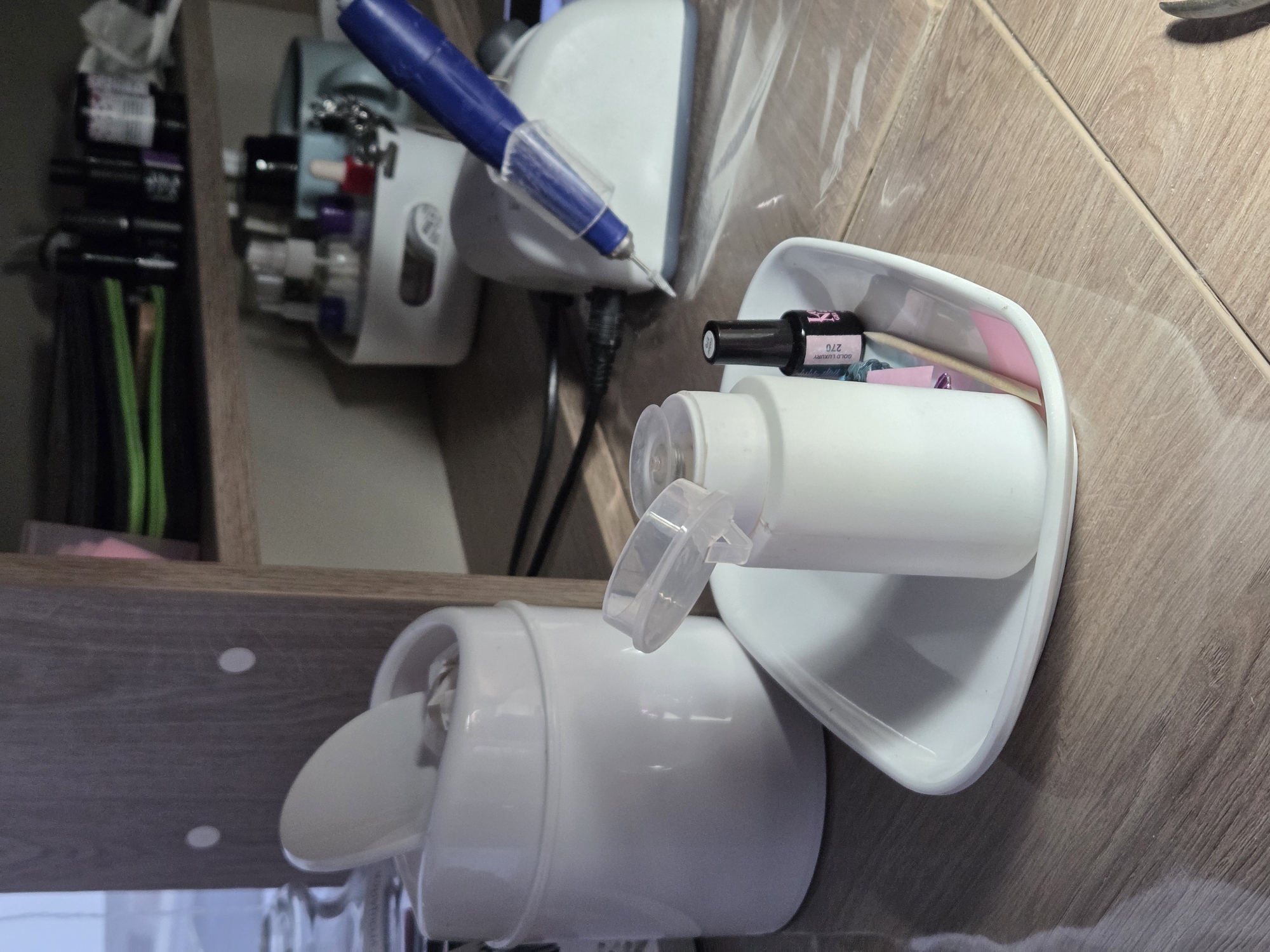 The image shows a tabletop with nail care items. A white nail drill with a blue handpiece is positioned on the left. Next to it, a white container with a hinged lid and another open container are present. The open container holds a bottle of black nail polish with the label "Gold Luxury 27G" and other small beauty tools. The surface is a light wood grain. The location is unknown.