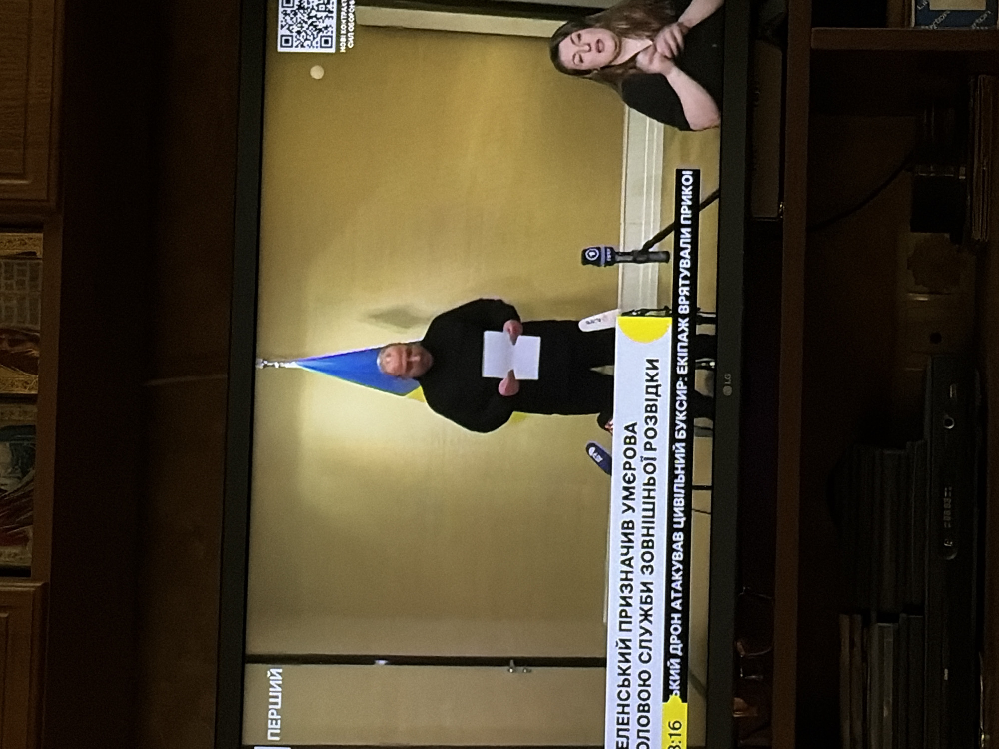 A man stands in front of a Ukrainian flag on a television screen, holding a paper. To his right, a woman is seated and gesturing with her hands, appearing to sign. A ticker tape at the bottom of the screen displays text in Ukrainian, and a QR code is visible on the upper right. The scene appears to be a news broadcast.
