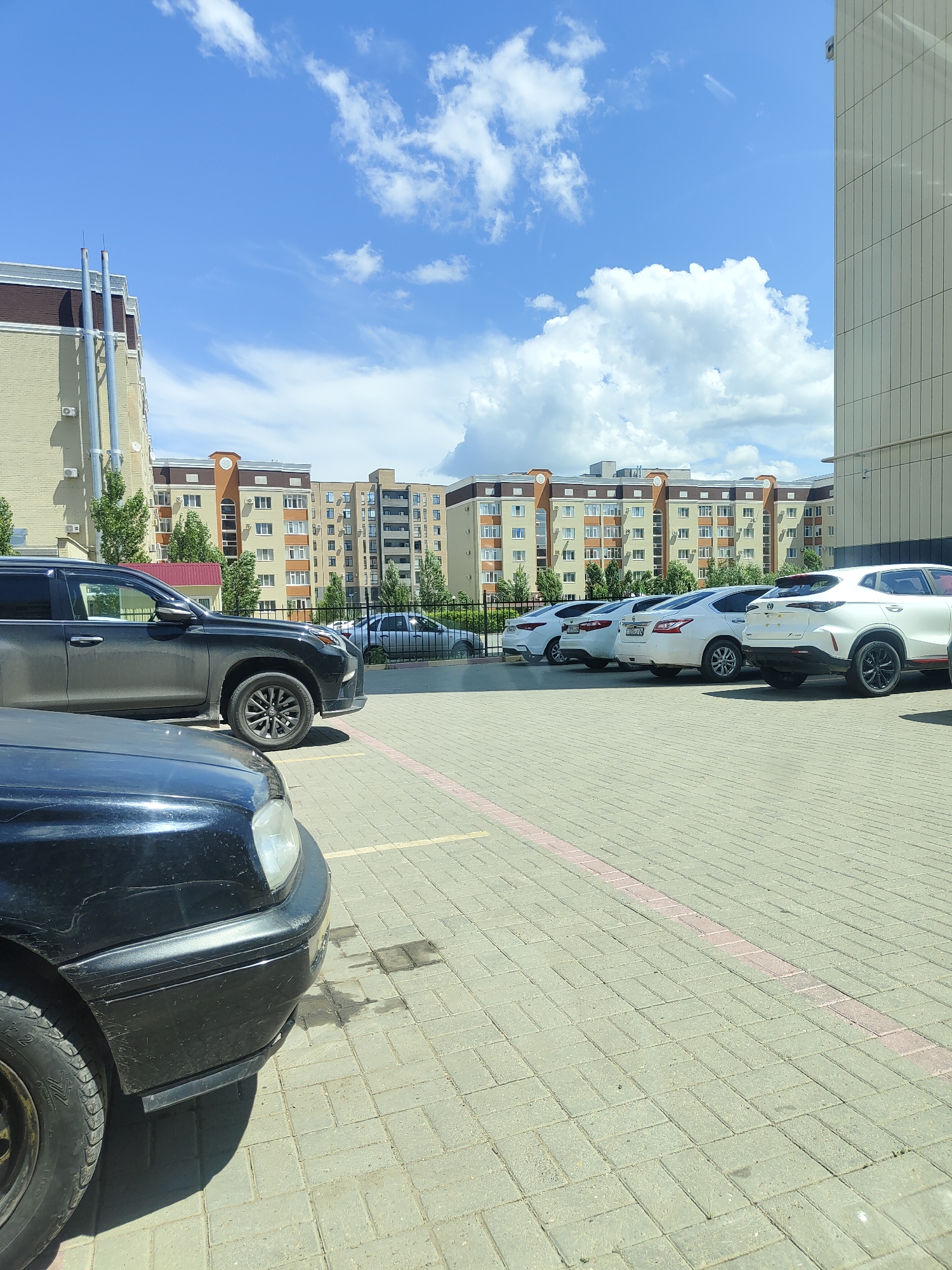 A bright, sunny day is captured in this exterior shot of a parking lot in Aktobe, Kazakhstan. The sky is a vibrant blue with scattered white clouds. The scene is dominated by several parked cars. In the foreground, the front of a black car is visible, showing its bumper and tire. To the right, a dark grey SUV is parked, with its side and wheels clearly in view. Further down the row, a line of white cars, including a white SUV on the far right, are parked.
Behind the parking lot, a modern residential building complex stretches across the background. The buildings are multi-story, with a light-colored facade and prominent dark brown and beige trim. Some of the buildings feature arched window designs and balconies. The architecture suggests a contemporary urban environment.
The parking lot itself is paved with light grey interlocking bricks, with a pinkish-red stripe marking the parking spaces. The overall impression is a typical residential parking area on a clear day. No people are visible in the frame. The time of day appears to be midday, given the strong sunlight and shadows. The location cues, such as the architectural style of the buildings and the presence of vehicles common in the region, along with the explicit mention of Aktobe, Kazakhstan, establish the setting.