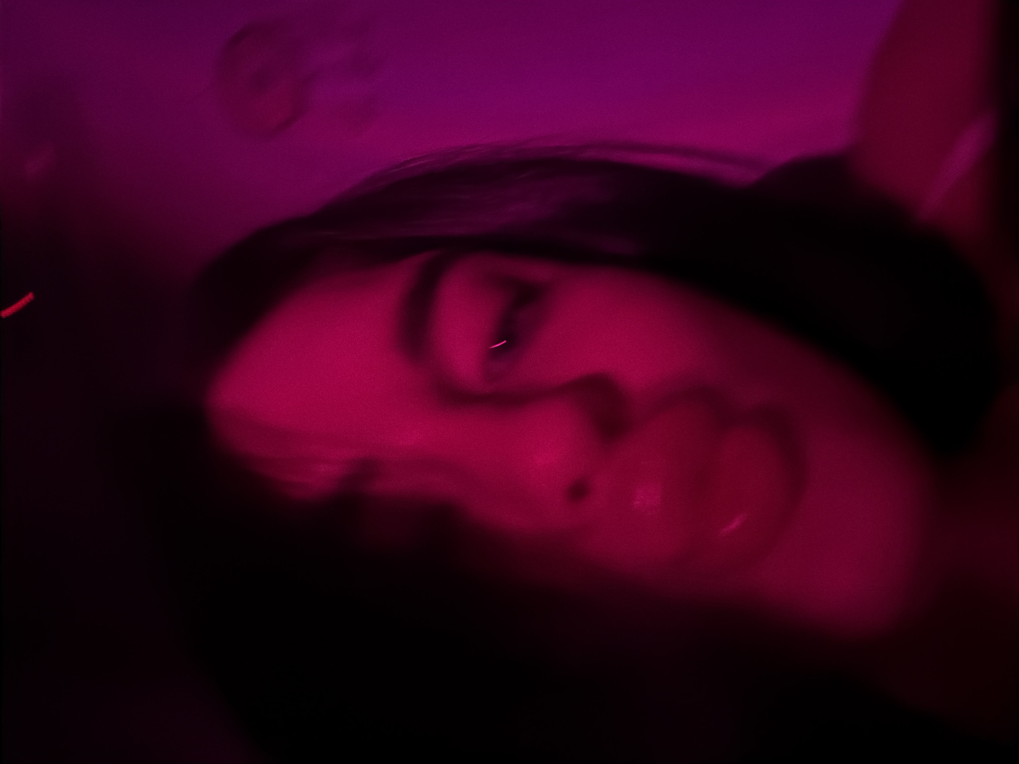 A face, likely a female, is prominently featured in the image, illuminated by a strong magenta-colored light. The face is slightly blurred. The hair appears dark. The background is mostly dark and indistinct. The image does not suggest any specific location, particularly Moscow, Russia.