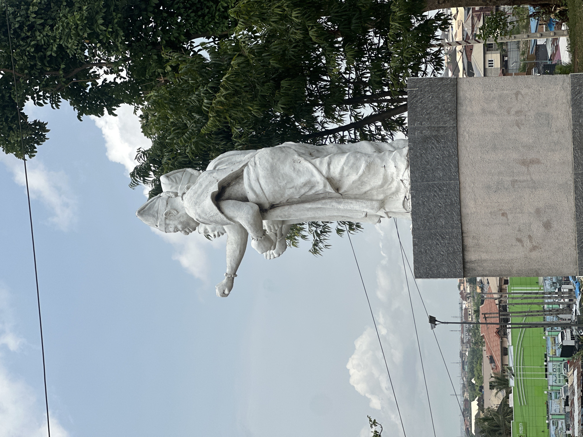 The image depicts a white marble statue of two figures, one supporting the other, situated in Ikeja, Nigeria. The statue stands on a rectangular gray stone base. Above and to the left of the statue are green leafy trees, against a backdrop of a blue sky with scattered white clouds. In the distance, buildings and infrastructure are visible, including a green-colored advertisement of ADORN. Electrical wires are strung across the scene.