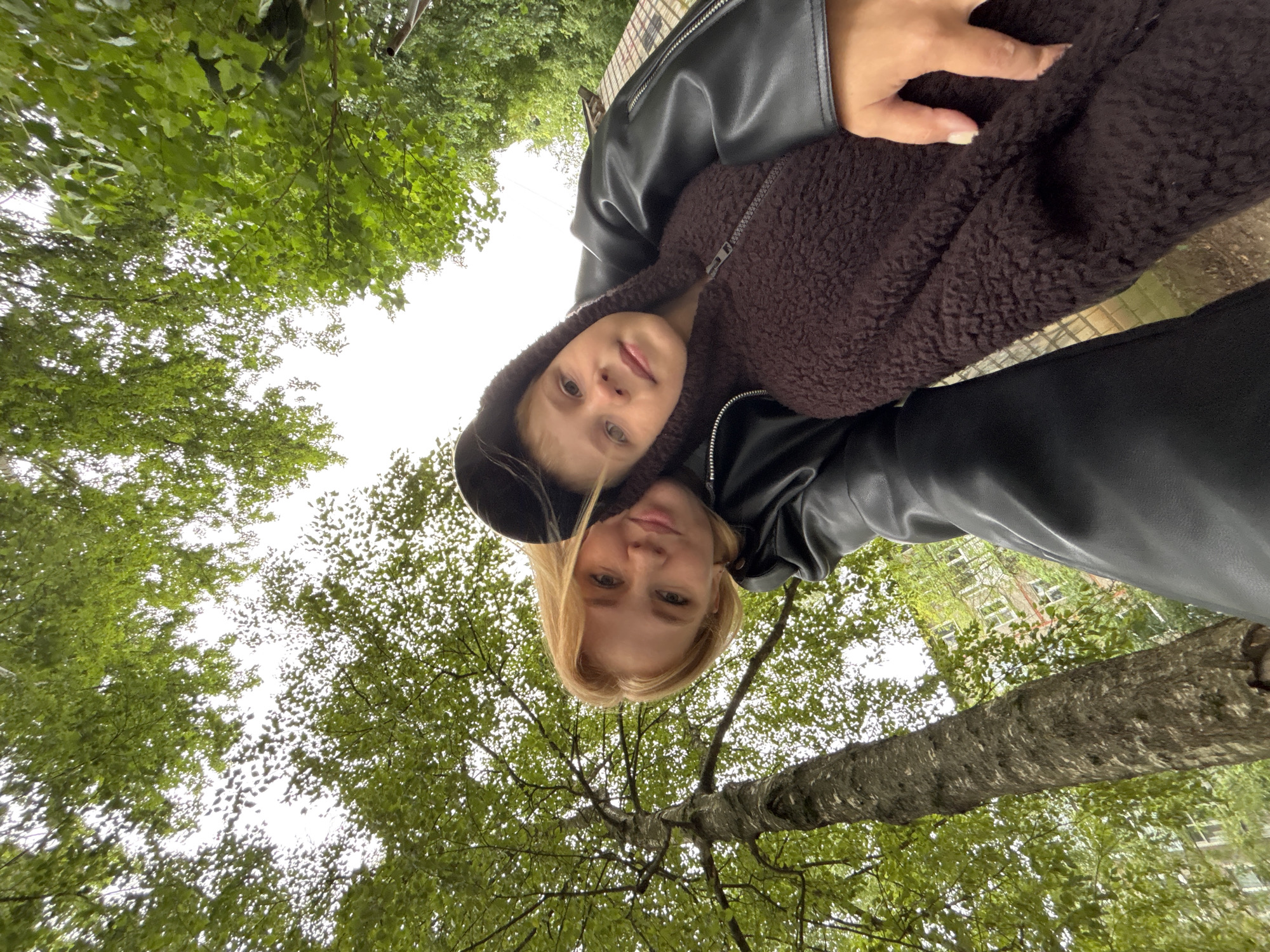 In Rusanovo, Russia, a low-angle, outdoor, medium shot captures a woman and a child posing for a selfie amidst a verdant, leafy environment. The woman, with blonde hair and a light complexion, is wearing a black leather jacket and is positioned on the left side of the frame. She is embracing the child, who is on the right, wearing a brown hooded jacket. The child's face is turned towards the camera, with light brown eyes and a gentle expression. The background is dominated by the lush green foliage of trees, with the sky visible through the leaves. A tree trunk, with textured bark, stands prominently on the left, and a building with windows can be seen in the distance on the right. The overall scene conveys a candid moment of connection between the two individuals.