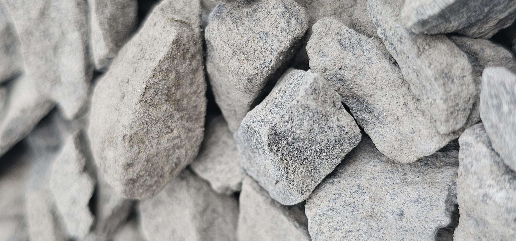 The image shows a close-up, macro view of a pile of gray, angular stones. The stones vary in size and shape, with some appearing rough and others smoother. They are covered in a fine, sandy dust, which gives them a matte appearance. The lighting is even, suggesting it might be daytime with overcast skies, or taken indoors with artificial lighting. There are no people, animals, or any other living beings visible. The setting is simply a collection of rocks, likely aggregate or gravel, with no discernible background or location cues beyond the texture and color of the stones. No text is visible in the image.