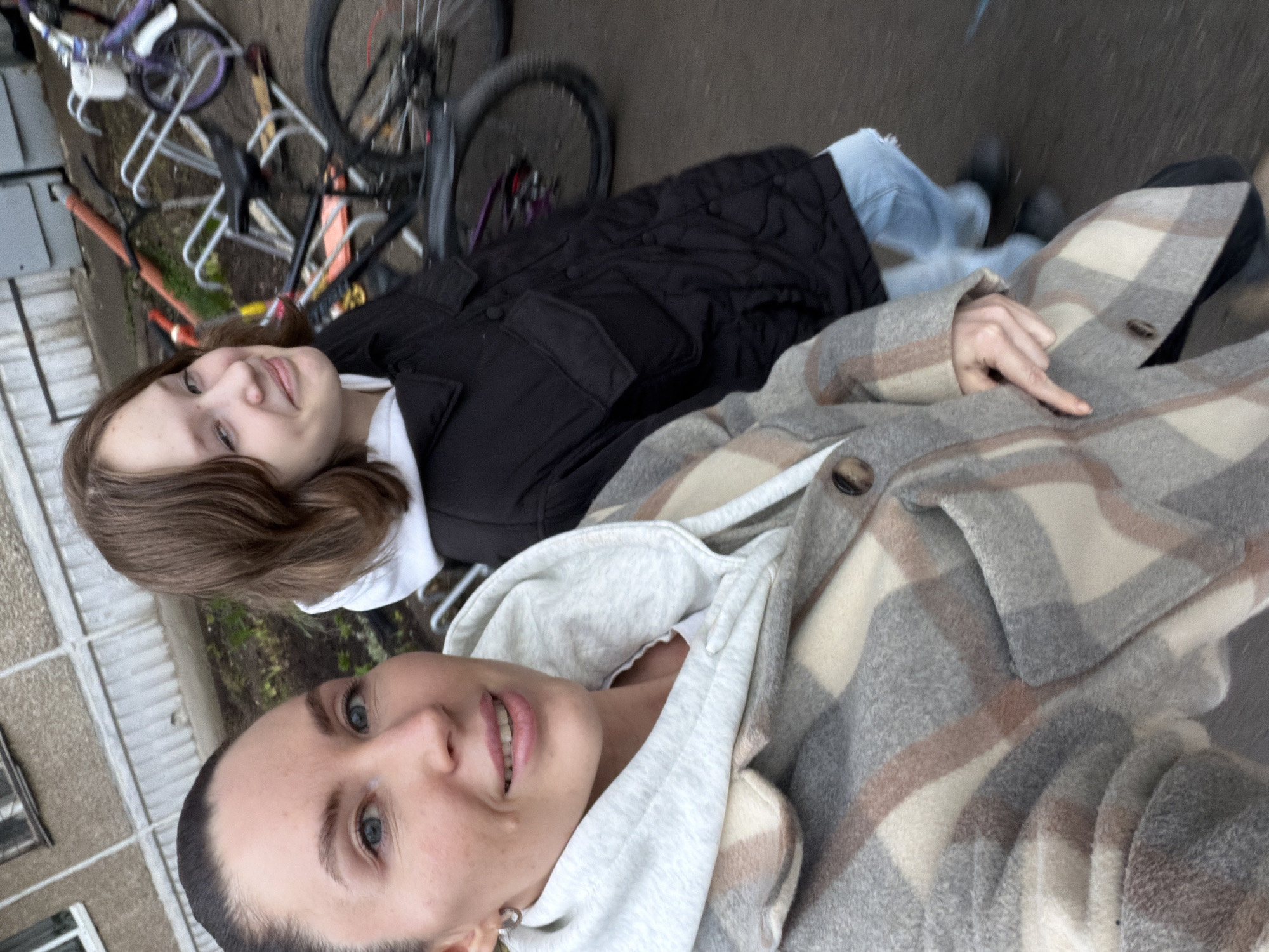Two females are photographed from a high angle, laying on a sidewalk. One female is positioned closer to the camera, with a light gray hooded sweatshirt and a gray plaid jacket with brown accents. The second female is positioned behind her, wearing a black puffy jacket over a white shirt and light blue jeans. A hand with light skin is extended, pointing downwards. A bicycle rack with bicycles are visible in the background, along with a gray building with windows. The location appears to be a city environment. Perm, Russia is given as the location. The sidewalk is gray with a hint of green.