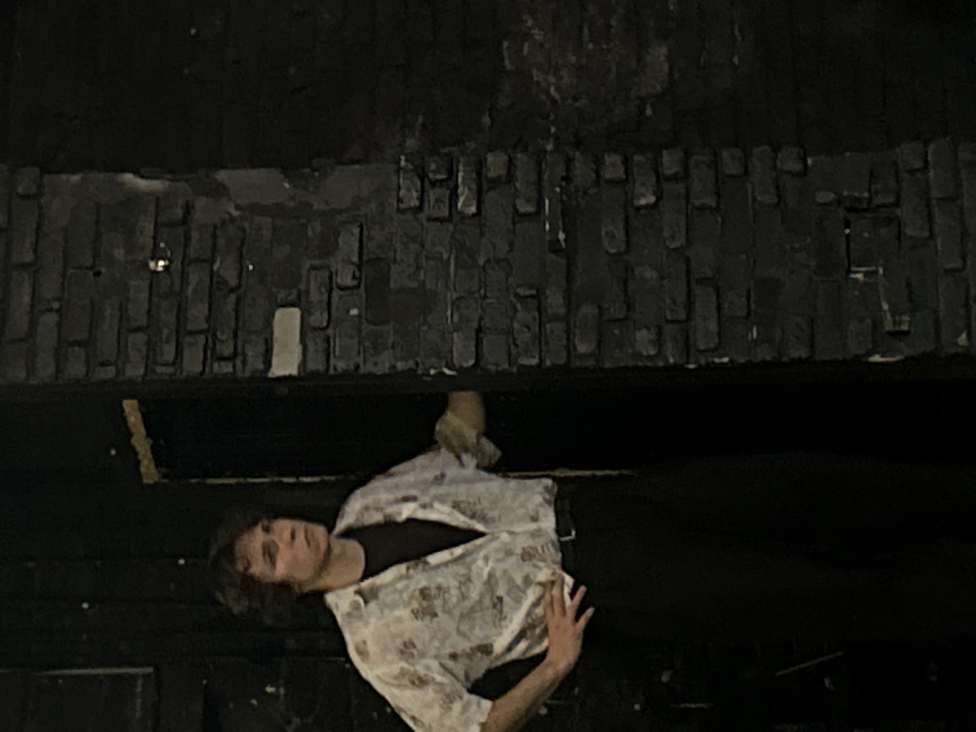 A person lies on their back, in what appears to be a dark, dimly lit space. They are wearing a black t-shirt and an unbuttoned, light-colored patterned shirt over it. Their legs are bent and their arms are resting on their body. The background consists of a rough, dark textured surface that resembles brickwork at the top of the image. Below the brickwork is a dark, flat surface. The lighting is low, casting shadows and obscuring details. The person's face is visible, with their head turned to their right, and they appear to be looking upwards. Their expression is neutral. The overall mood is somber and still, suggesting a moment of rest or contemplation. The image is taken from a high angle, looking down on the subject. There is no visible text in the image.