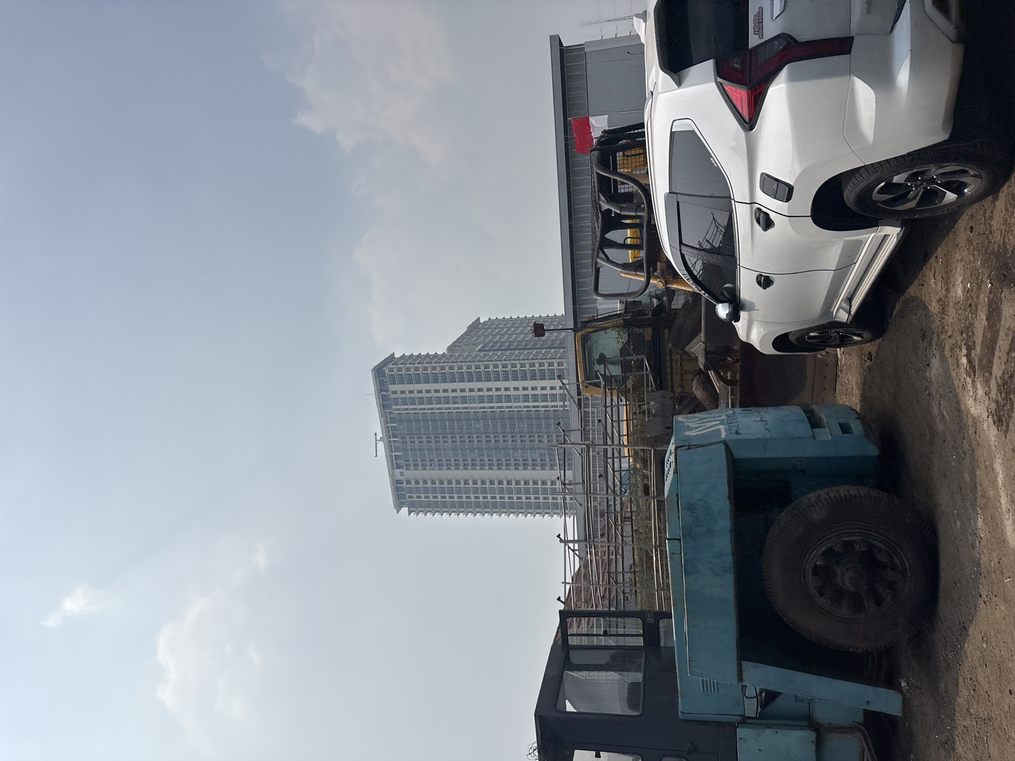 The image depicts an outdoor scene in Bekasi, Indonesia, characterized by a construction or industrial area. In the foreground, a white SUV is parked on a dirt surface. To the left of the SUV, a large, teal-colored industrial vehicle, possibly a forklift or tow truck, is visible, its front tire prominent. Behind these vehicles, a yellow excavator is partially visible. Further back, a modern, multi-story building with a light-colored facade stands tall against a hazy sky. Adjacent to the excavator and in front of the tall building is a low-rise industrial building with a metal facade. A small Indonesian flag, red and white, is mounted on the roof of this industrial building. The sky is overcast with some clouds. The overall impression is one of ongoing development or operation within an urban industrial setting in Bekasi.