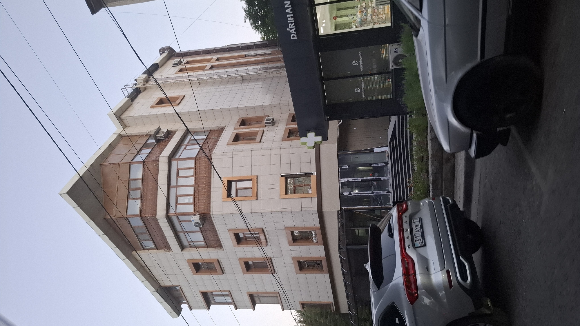 The image captures a modern, multi-story building in Shymkent, Kazakhstan, with a pharmacy on the ground floor. The building features a beige tiled facade with numerous rectangular windows, some with brown trim and decorative ironwork on the lower levels. A distinctive angled section with a copper-colored paneling and recessed windows protrudes from the upper floors. The pharmacy, branded with a green cross and the text "DARİHAN," has large glass windows displaying shelves stocked with pharmaceutical products. Two cars are visible on the street in front of the building: a silver SUV on the left and the front portion of a silver sedan on the right. Overhead power lines crisscross the sky, indicating an urban setting. The sky is clear and appears to be either early morning or late evening due to the soft lighting. The overall scene suggests a commercial area within the city.