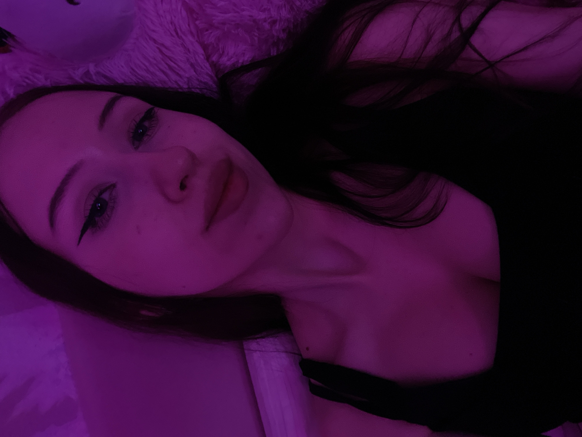 The image shows a close-up, selfie-style shot of a young woman illuminated by purple light. She has dark hair, winged eyeliner, and is wearing a black tank top. The background appears to be a soft, textured surface, possibly a blanket or pillow, also bathed in the purple light. The scene is a personal, indoor setting. There is no indication of the city of Votkinsk, Russia, or any specific location within it.