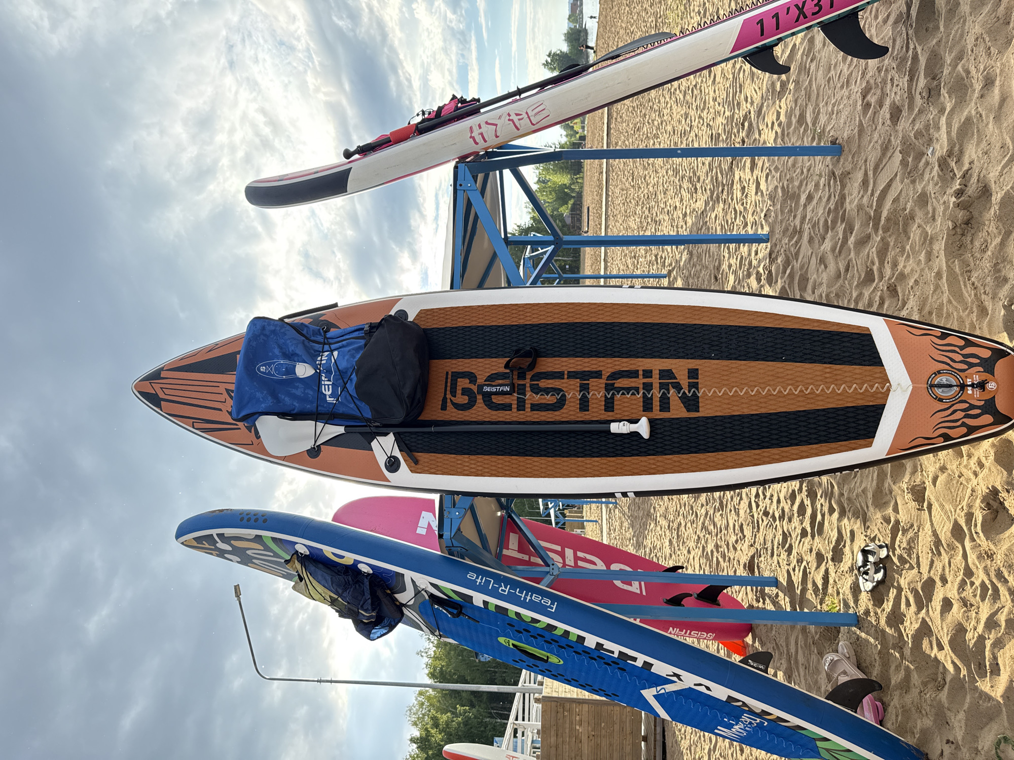 The image depicts a collection of stand-up paddleboards (SUPs) resting on a sandy beach. The scene is set outdoors under a partly cloudy sky, suggesting it could be daytime. The paddleboards are arranged on a blue metal rack, with some lying flat on the sand.
The foreground features a large, orange and black striped paddleboard with "BEISTEIN" prominently displayed in black lettering. A blue bag is placed on top of this board. Above it, another paddleboard, primarily white with pink and black accents, is visible, bearing the text "HYFE" and "11'X31". Further down, a pink paddleboard is partially obscured by a blue paddleboard in the lower portion of the image. This blue board has "Feath-R-Life" written on it in white lettering.
The sand shows the typical texture of a beach, with footprints and ripples. In the background, trees and a body of water, likely a lake or river, can be seen, suggesting a recreational area. There are no people clearly visible in the immediate foreground or interacting with the paddleboards, implying they might be stored or waiting to be used. The overall impression is a tranquil beach setting, possibly during a summer day, ready for water activities. The orientation of the image is portrait, with the subjects appearing tilted.