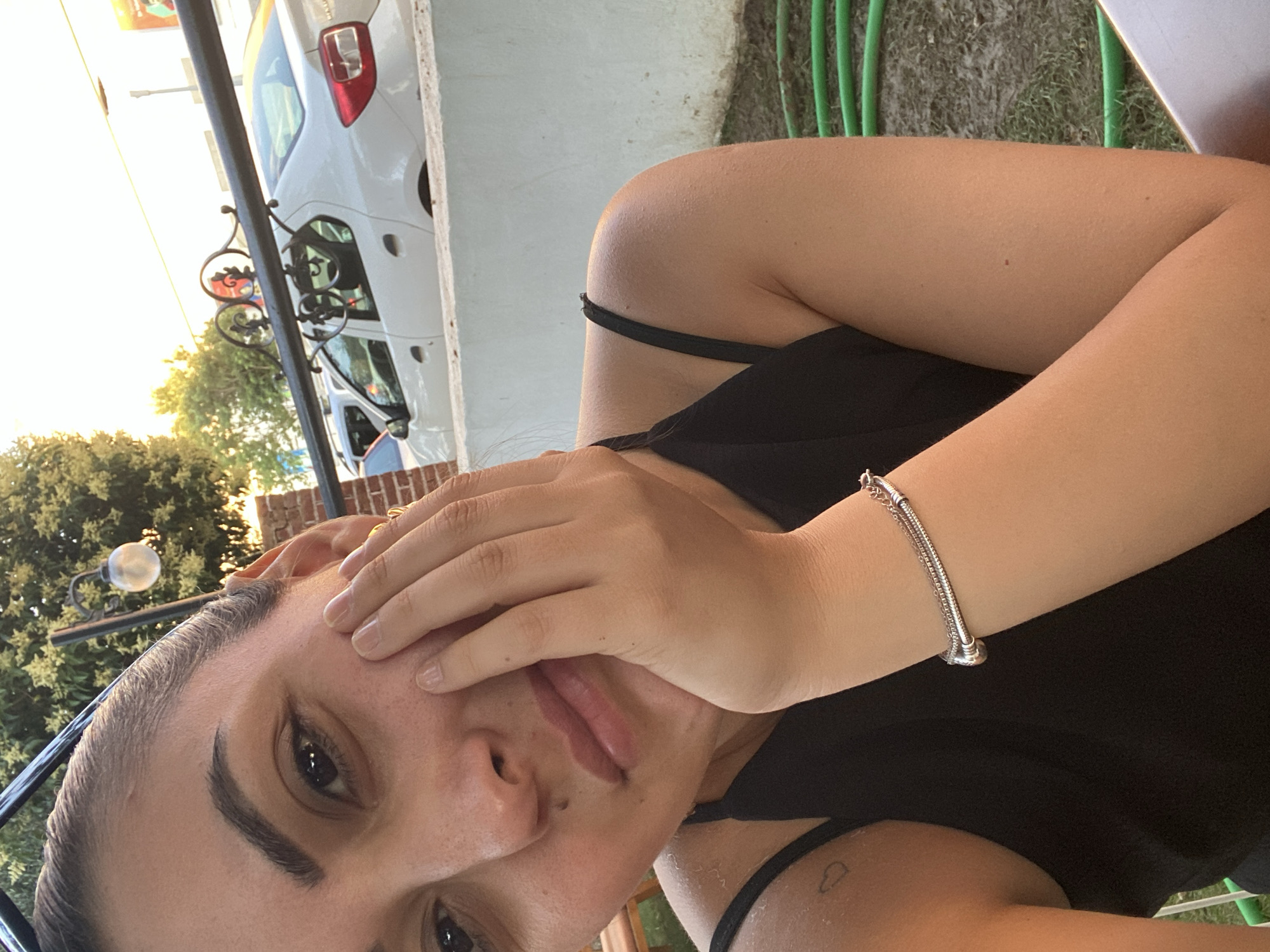 The image shows a close-up, selfie-style shot of a woman with her hand resting on her cheek. She has dark hair pulled back, dark eyes, and light skin with some freckles. She is wearing a black tank top and a silver bracelet on her right wrist. Behind her, a white car is visible parked on a street. The background also includes trees, a brick wall, and a decorative metal railing. The lighting suggests it is late afternoon or early evening. The image appears to have been taken outdoors, possibly in a residential or public area of Yunus Emre, Turkey, given the context.