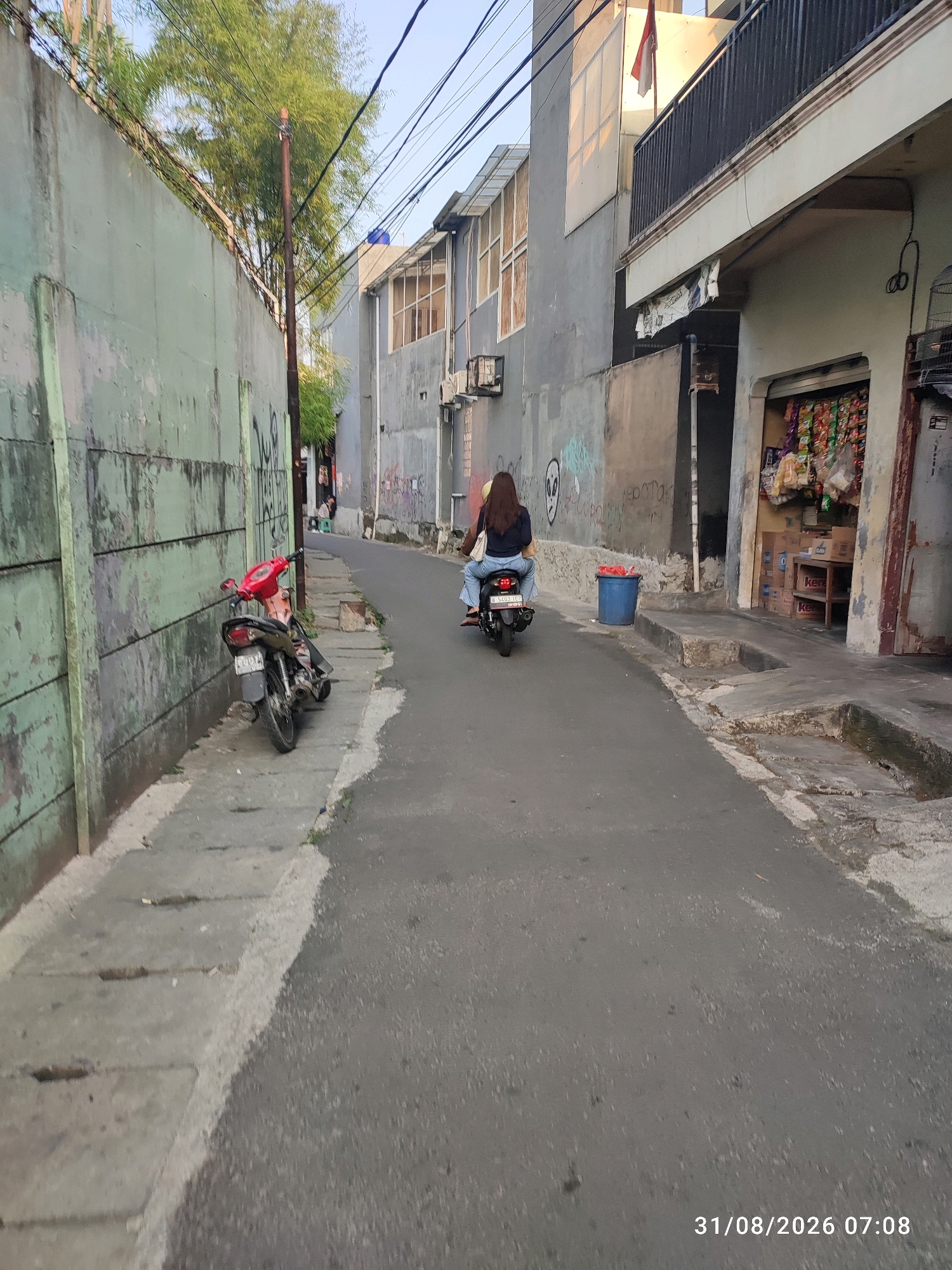 The image depicts a narrow street scene in East Jakarta, Indonesia, with buildings lining both sides. On the left, a weathered, pale green concrete wall runs alongside the street, marked with graffiti. A red and black motorcycle is parked on the gray concrete sidewalk next to the wall. The street itself is paved with asphalt.
On the right side of the street, a building with a balcony displays an Indonesian flag. Below the balcony, a small shop is visible, with shelves stocked with various bagged snacks and drinks. A blue trash can is placed on the sidewalk near the shop.
A woman is riding a black motorcycle down the street, with her back to the camera. She is wearing a dark top and blue jeans, and she appears to be carrying a light-colored bag on her shoulder. The time stamp "31/08/2026 07:08" is present in the lower right corner of the image. Overhead, a tangle of electrical wires crisscrosses the sky, with some leafy green trees visible in the background, suggesting an urban environment with pockets of vegetation.