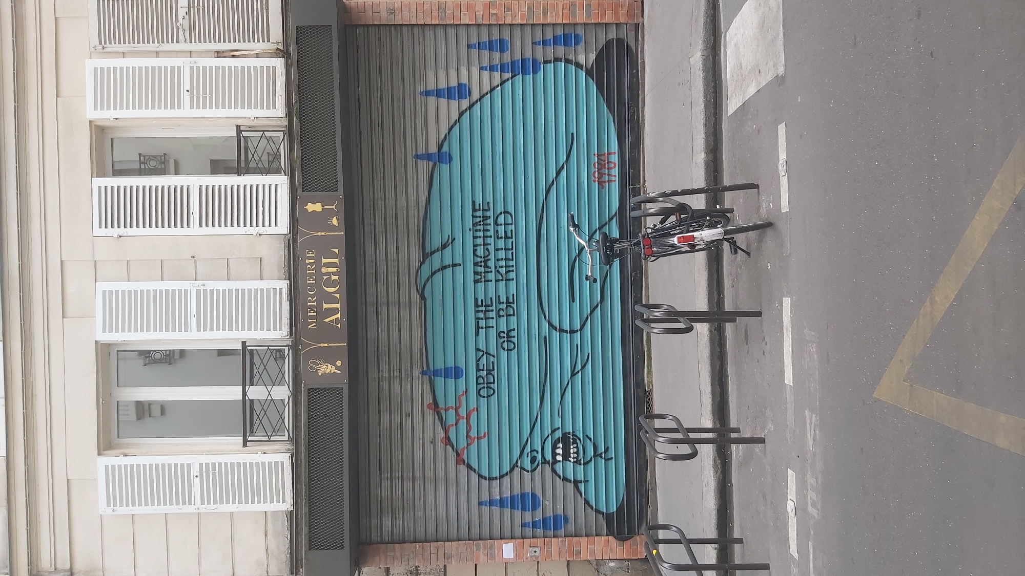 A graffitied metal roll-up door on a building in Paris, France, features a large, teal-colored illustration of a stylized, grotesque face. The face is rendered with a gaping mouth, sharp teeth, and red, veiny lines on its forehead, suggesting pain or distress. Blue teardrop shapes are depicted around the face, and the words "OBEY THE MACHINE OR BE KILLED" are spray-painted across it in black lettering. To the right of the graffiti, the year "1984" is written in red. Above the roll-up door, a sign for "MERCERIE ALGIL" is visible, adorned with illustrations of sewing tools. The building has multiple windows with white shutters, and the street in front of the shop has a bicycle rack with a bicycle parked in it.