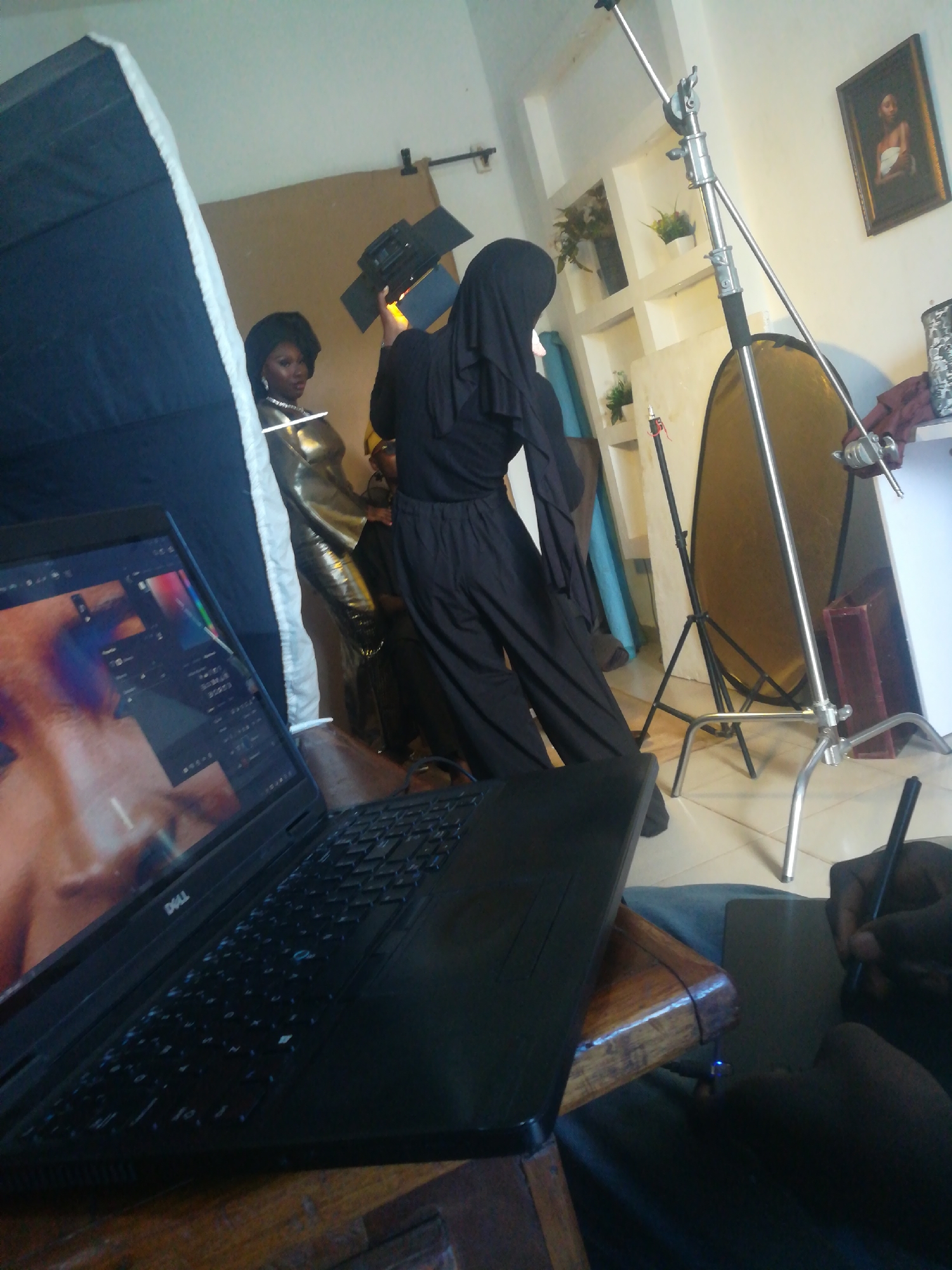 In an indoor studio setting, two women are the subjects of a photo shoot. One woman, on the left, wears a gold, metallic-looking dress and has dark hair. The other woman, on the right, is dressed in all black, including a hijab. She holds a lighting apparatus.
In the foreground, a black Dell laptop is open on a wooden table, displaying an image that appears to be part of the photo shoot. A person's hand is visible on the right, holding a stylus over a black tablet or another device.
The background of the studio shows various photography equipment, including a light stand, a large collapsible reflector, and what appears to be a backdrop or fabric. There are also shelves on the wall with decorative items and a framed picture. The overall environment suggests a professional or semi-professional photography setup.