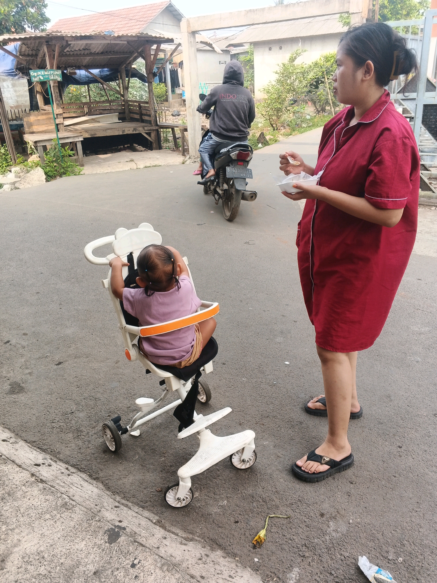 In Kadu, Indonesia, a woman in a red robe stands on an asphalt road, holding a white bowl and a spoon, with a toddler seated in a white stroller. The toddler faces away from the camera, wearing a purple shirt and beige shorts. A person on a motorcycle, wearing a grey hoodie, rides away from them. In the background, a wooden structure with a corrugated metal roof is visible, along with buildings and vegetation. The scene appears to be a residential street.
