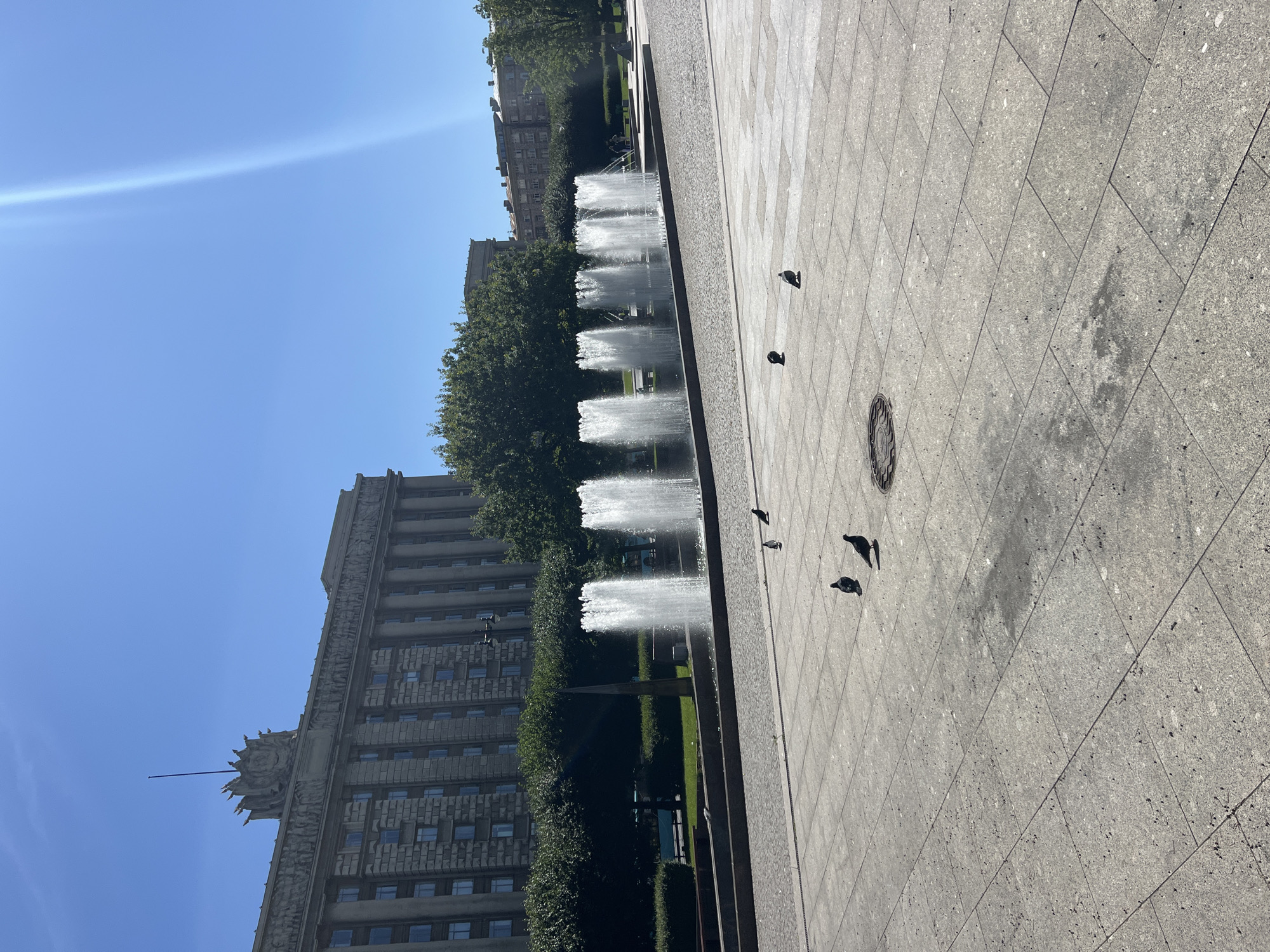 This is a photograph of the Alexander Nevsky Square in Saint Petersburg, Russia. The scene depicts a wide, paved plaza with several pigeons scattered across the ground. In the background, a large, neoclassical building with columns and ornate architectural details stands under a clear blue sky. To the left of the building, a row of trees forms a green barrier. In the middle ground, a series of fountains are active, sending jets of water into the air. The sun is bright, casting shadows and creating a lens flare in the upper right portion of the image.
