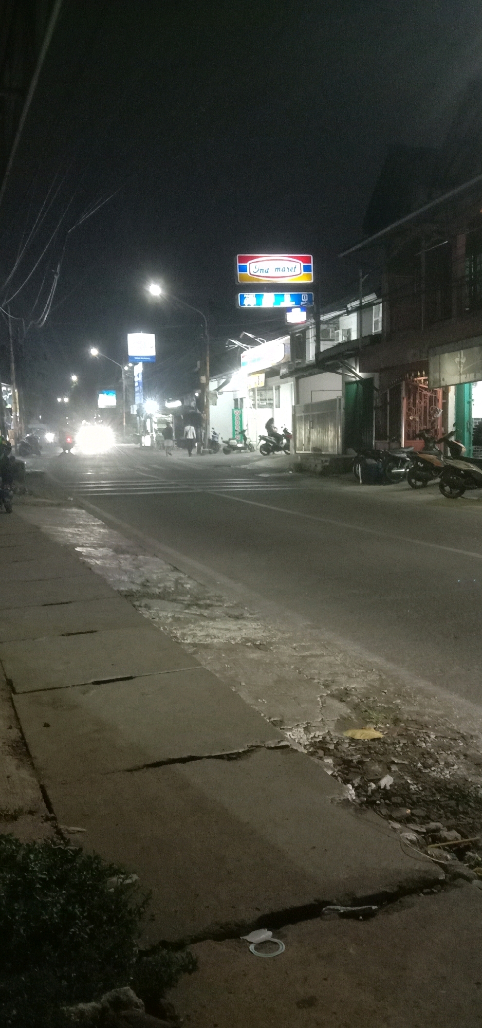 The image depicts a street scene at night in Tangerang, Indonesia, with a prominent "Indomaret" convenience store sign illuminated above a building. The street is paved, and a sidewalk runs along the left side of the road, showing signs of disrepair with cracked concrete and debris. Several motorcycles are parked on the right side of the street, and a few people are walking in the middle of the road. Headlights from oncoming vehicles create streaks of light. The overall atmosphere is urban and dimly lit, typical of a residential or commercial street after dark.